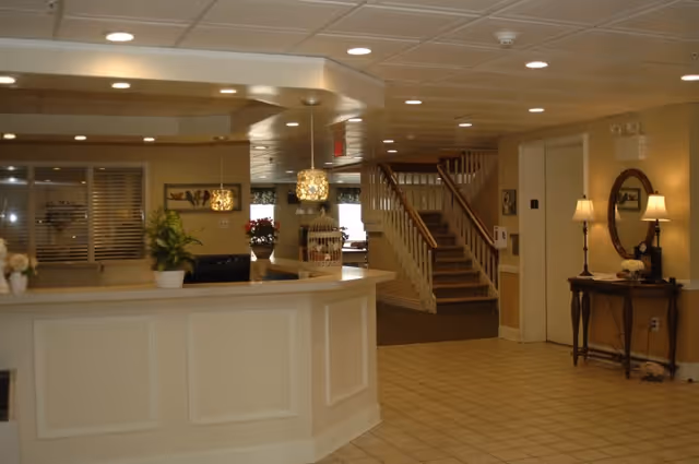 Interior view of a senior living facility reception area with a white front desk, potted plants, hanging pendant lights, a staircase leading upstairs, and a small table with a mirror and two lamps on the right side.