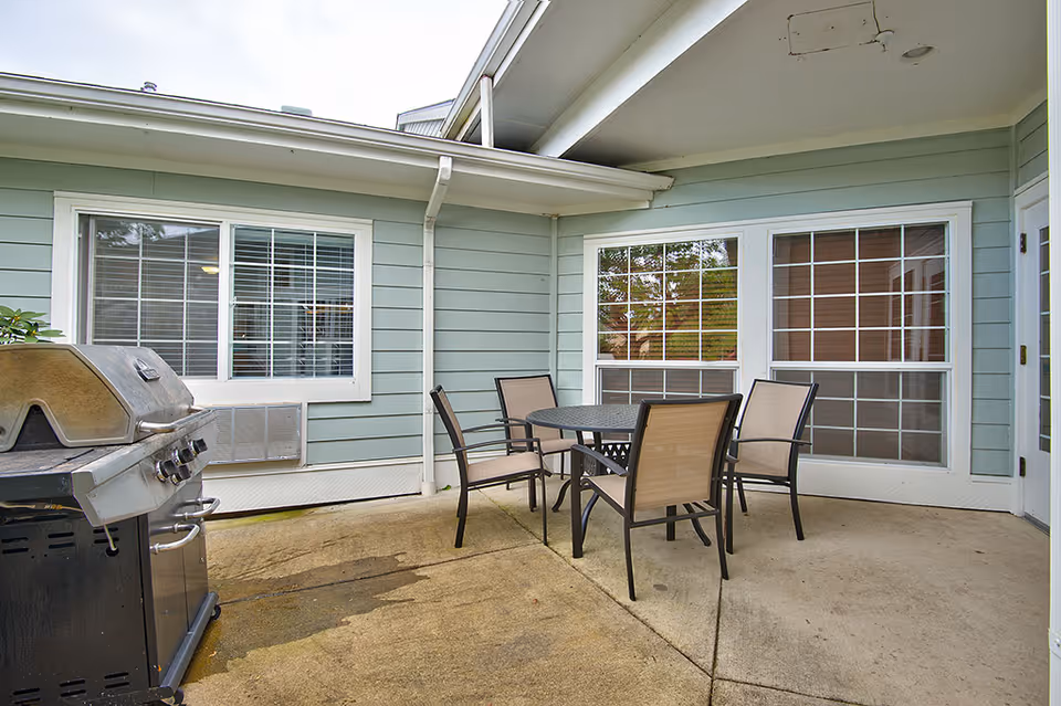 Outdoor patio area with a round metal table and four beige mesh chairs. There is a gas grill on the left side and light blue siding with white trim on the building walls surrounding the patio. Two large windows and a door are visible on the building walls.