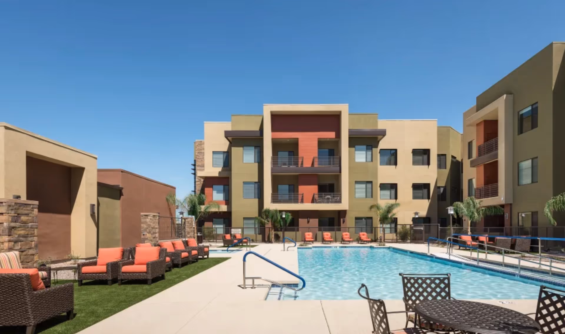 Outdoor swimming pool area at Clearwater Ahwatukee facility with lounge chairs featuring orange cushions, palm trees, and a modern three-story building in the background under a clear blue sky.