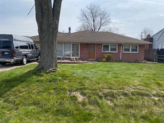 Single-story brick residential building with a front lawn and a large tree in the yard. A driveway on the left side has a black van parked. The sky is clear with some clouds.