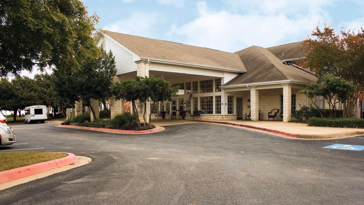 Exterior view of Brookdale Round Rock facility showing the main entrance with a covered drop-off area, surrounded by trees and landscaping, with a parking lot in the foreground.