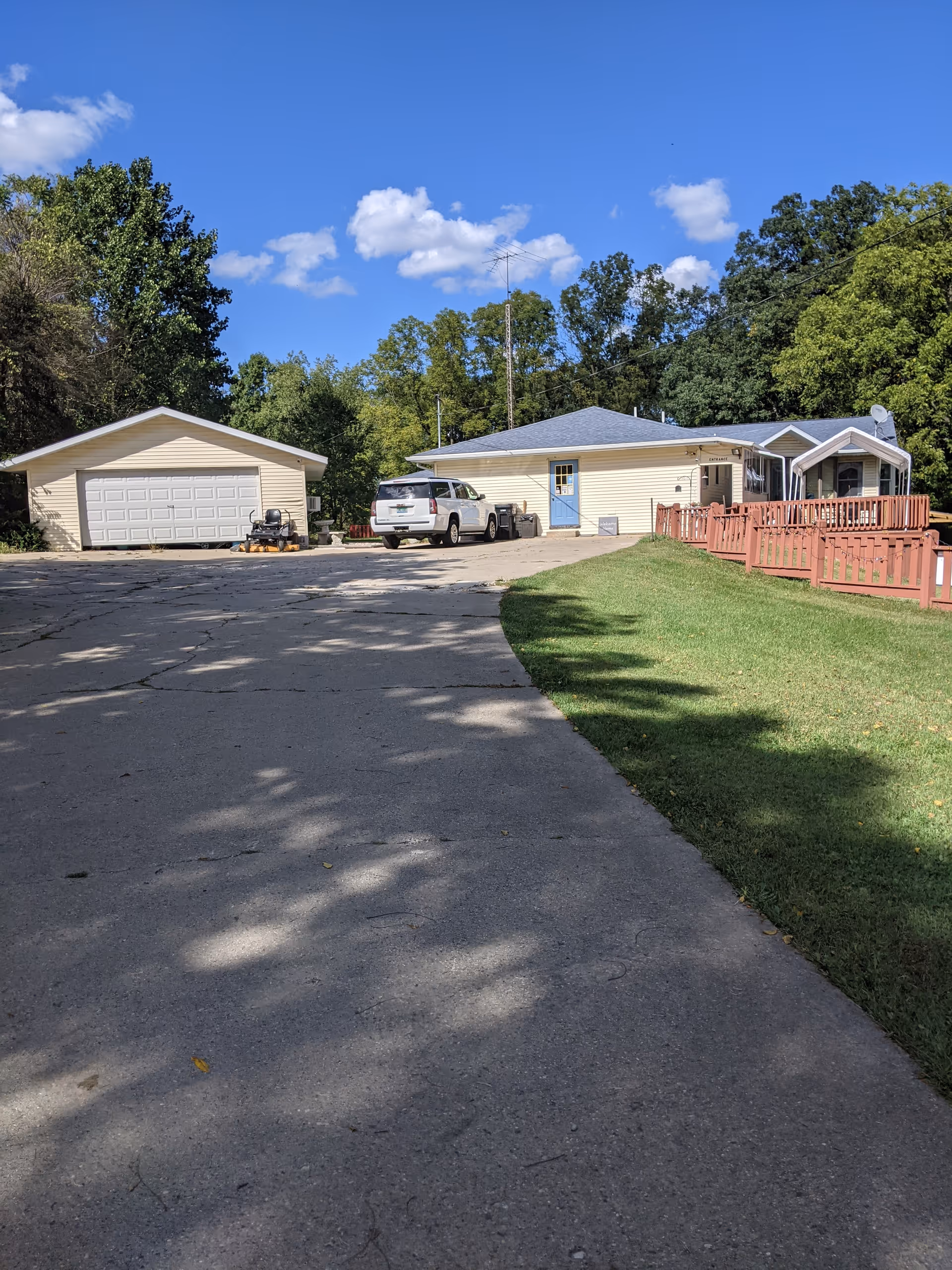 A driveway leading up to a single-story building with a blue door and a detached garage. There is a white SUV parked near the building. The area is surrounded by green trees and a clear blue sky with some clouds. A red wooden fence runs along the right side of the driveway.