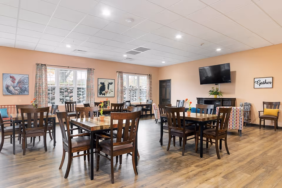 A spacious dining room in a senior living facility with multiple wooden tables and chairs arranged neatly. The room has light peach-colored walls, large windows with patterned curtains, and framed artwork of farm animals on the walls. A flat-screen TV is mounted on one wall above a dark wooden cabinet, and there are decorative flowers on the tables and cabinet. The floor is wood laminate, and the ceiling has recessed lighting.