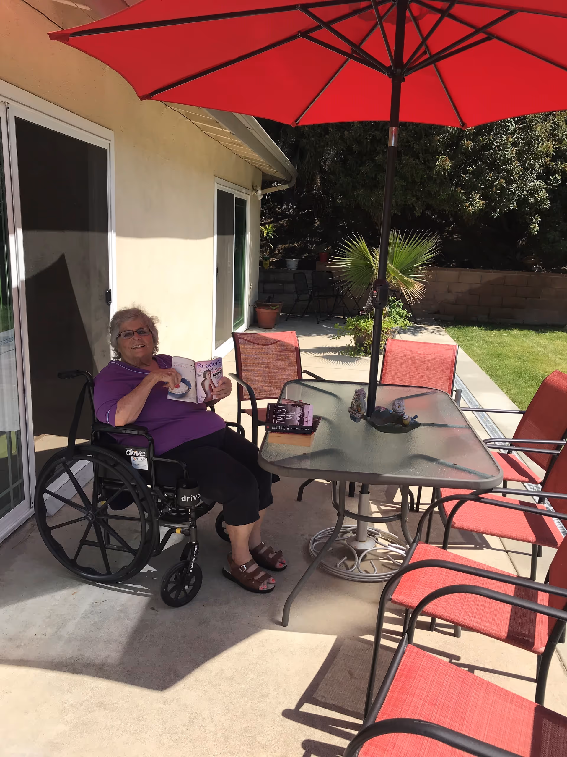 An elderly woman in a wheelchair sitting on a patio next to a glass table with red chairs and a large red umbrella. She is holding a book and smiling. The patio is adjacent to a house with sliding glass doors and there is greenery and a lawn in the background.
