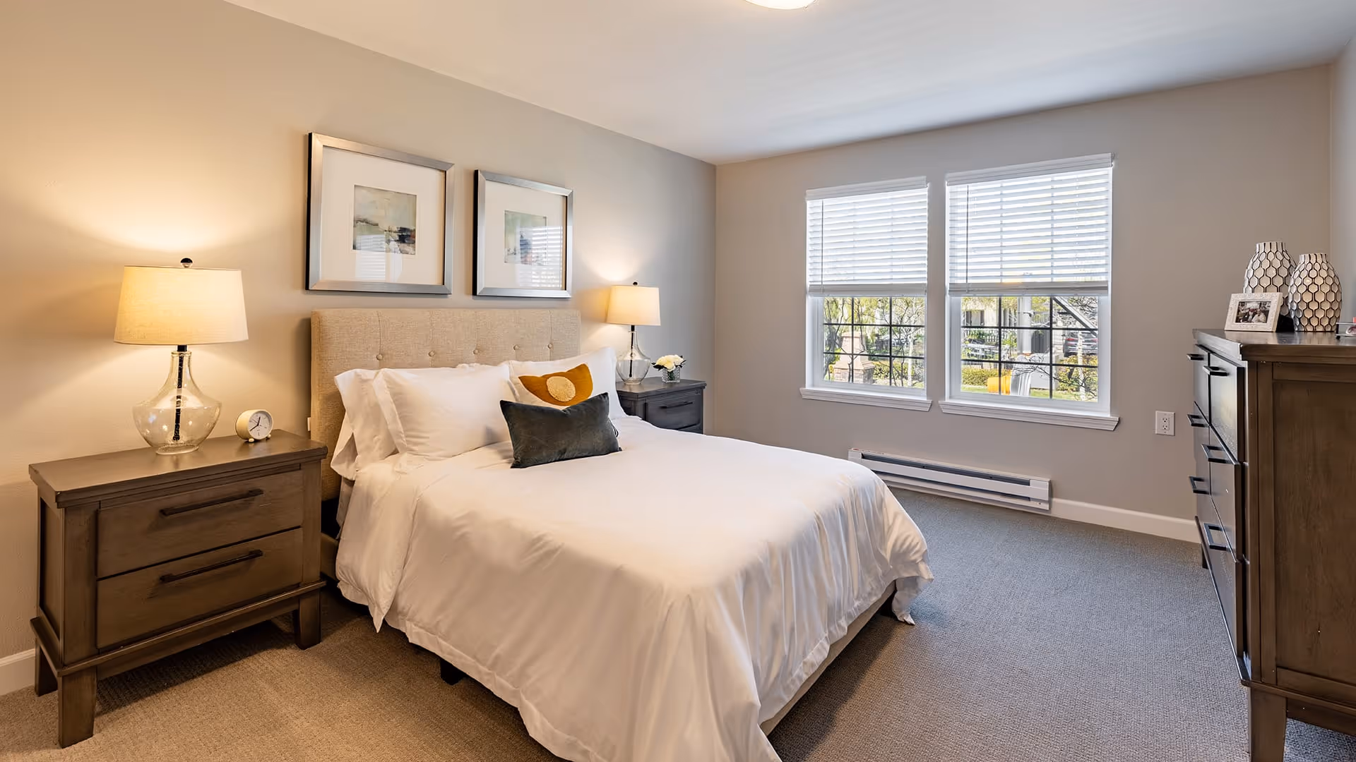 A neatly made bed with white linens and decorative pillows in a bedroom. The room features two wooden nightstands with lamps, two framed pictures above the bed, a window with blinds letting in natural light, and a wooden dresser with decorative items on top.