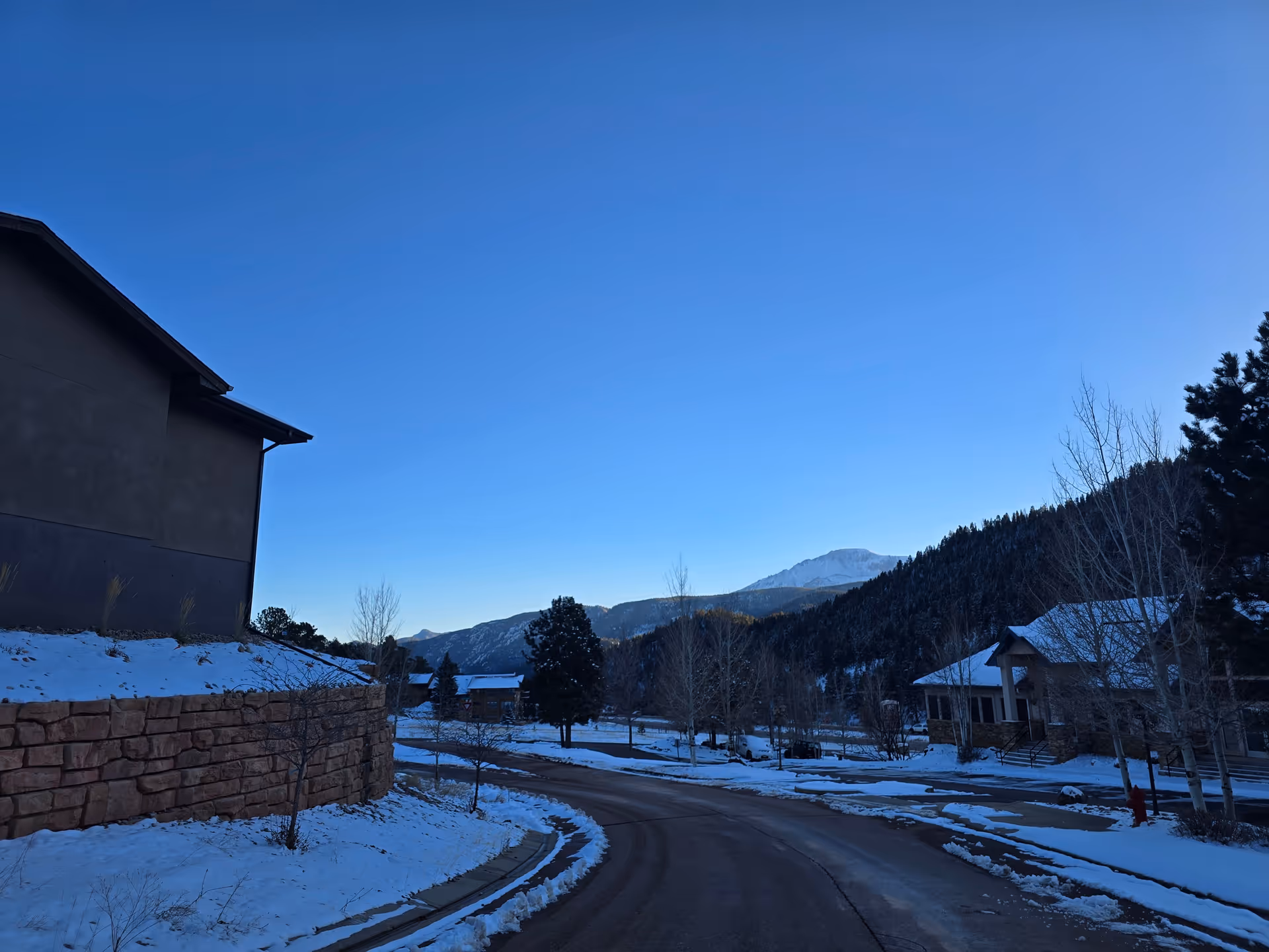 Snow-covered residential street with houses, trees and mountains under a clear blue sky.
