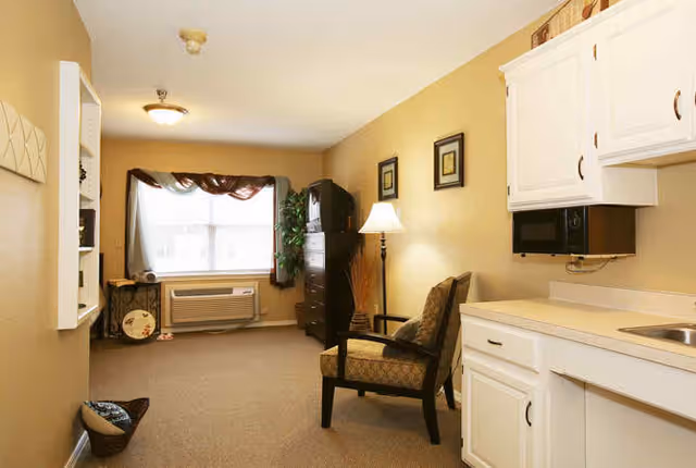 Interior view of a senior living facility room at Broadmoor Court featuring beige walls, a window with a valance, an air conditioning unit below the window, a dark wooden dresser with a TV on top, a floor lamp beside an upholstered armchair, and a kitchenette area with white cabinets, a microwave, and a sink.
