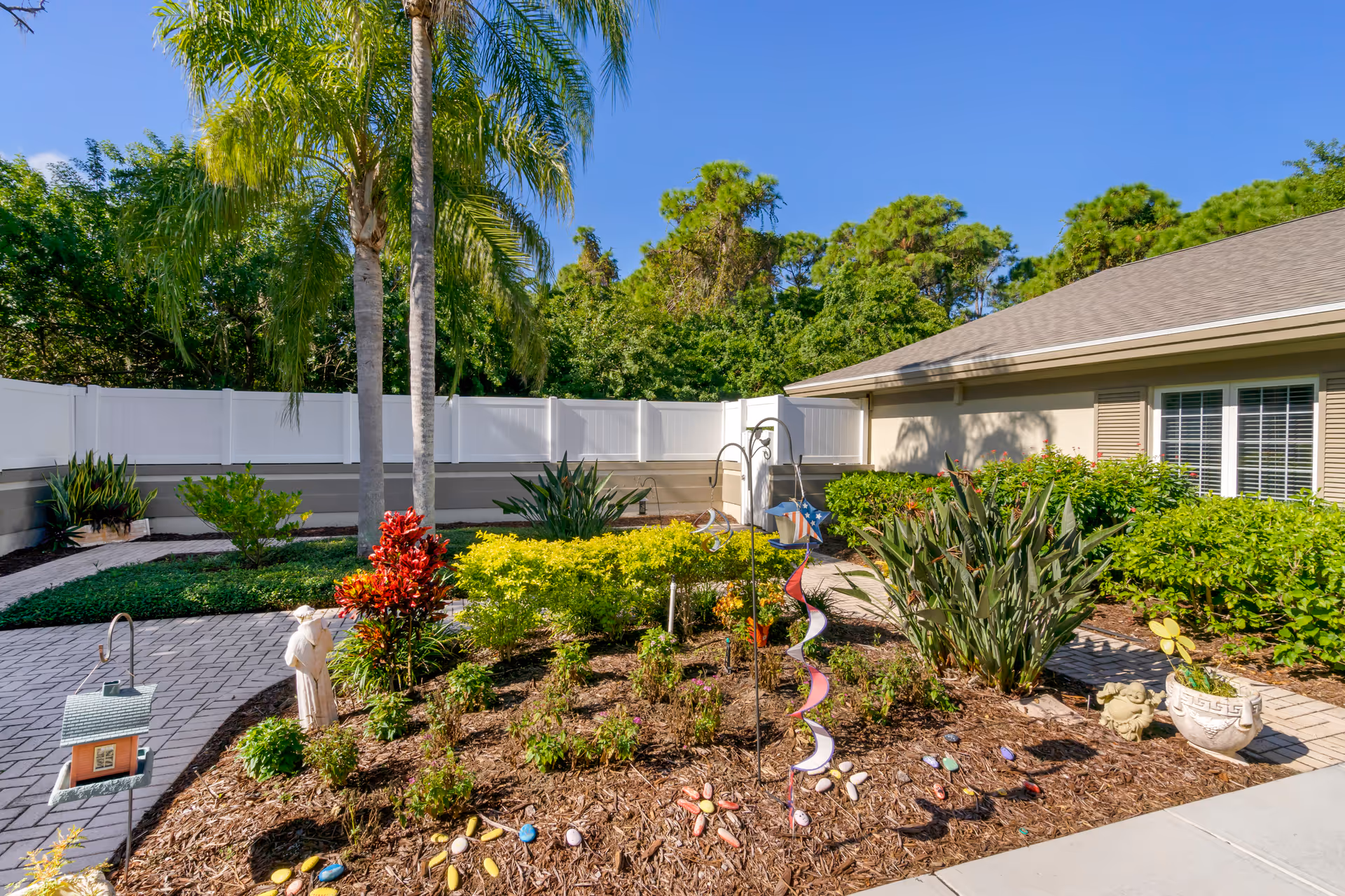 Sunny landscaped courtyard with palm trees, flower beds, decorative garden ornaments and a paved walkway beside a single-story building.