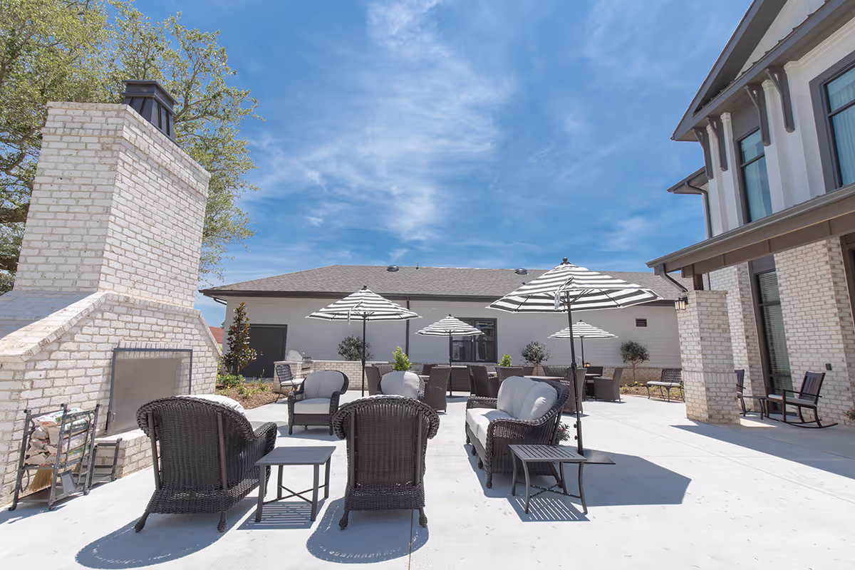 Outdoor patio with wicker seating, striped umbrellas, and a brick fireplace beside a facility building under a blue sky.