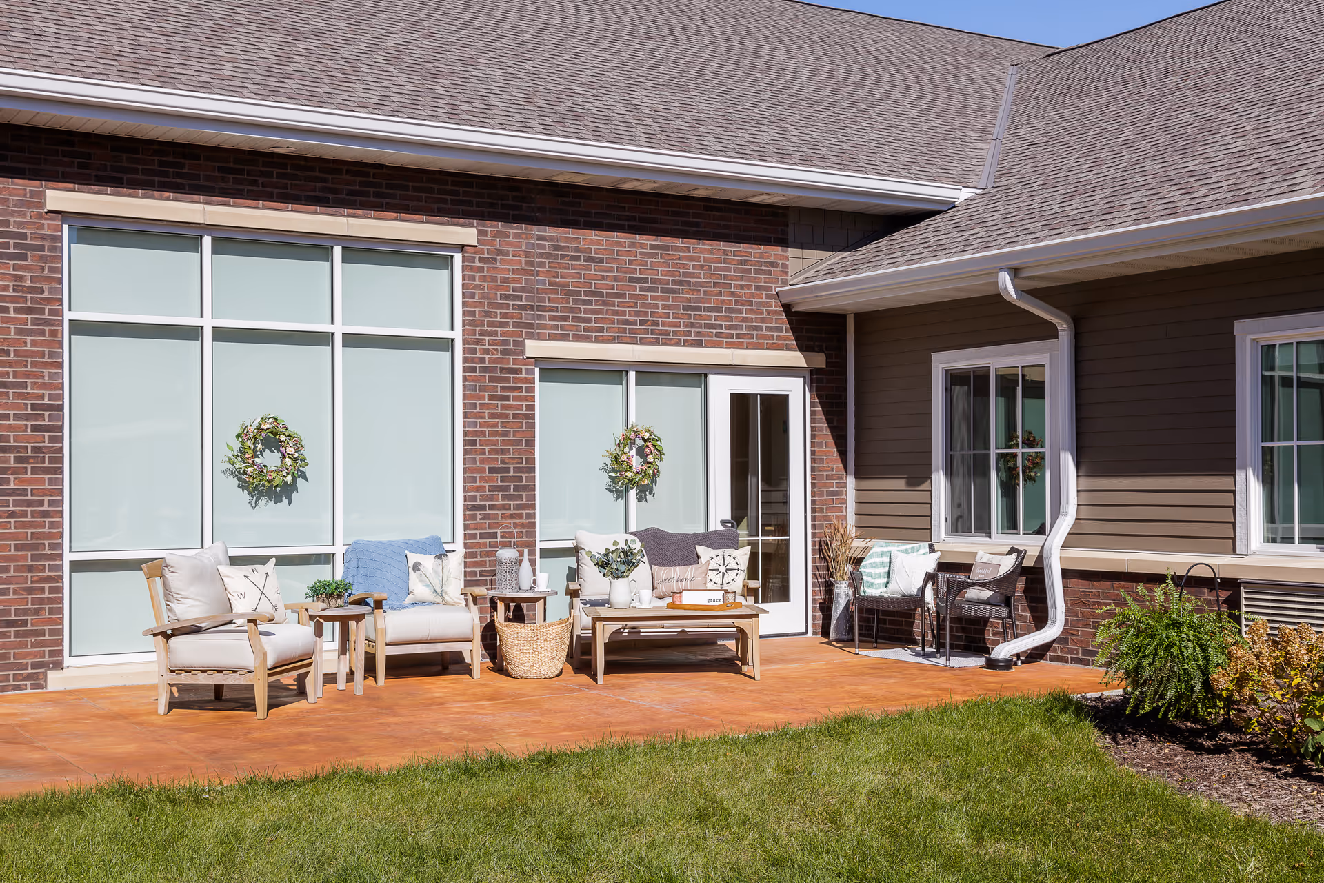Outdoor patio area at Edencrest at Siena Hills featuring cushioned wooden chairs, a small wooden table, a bench with pillows, and decorative wreaths on the windows. The patio is adjacent to a brick and siding building with a grassy lawn and some plants in the foreground.