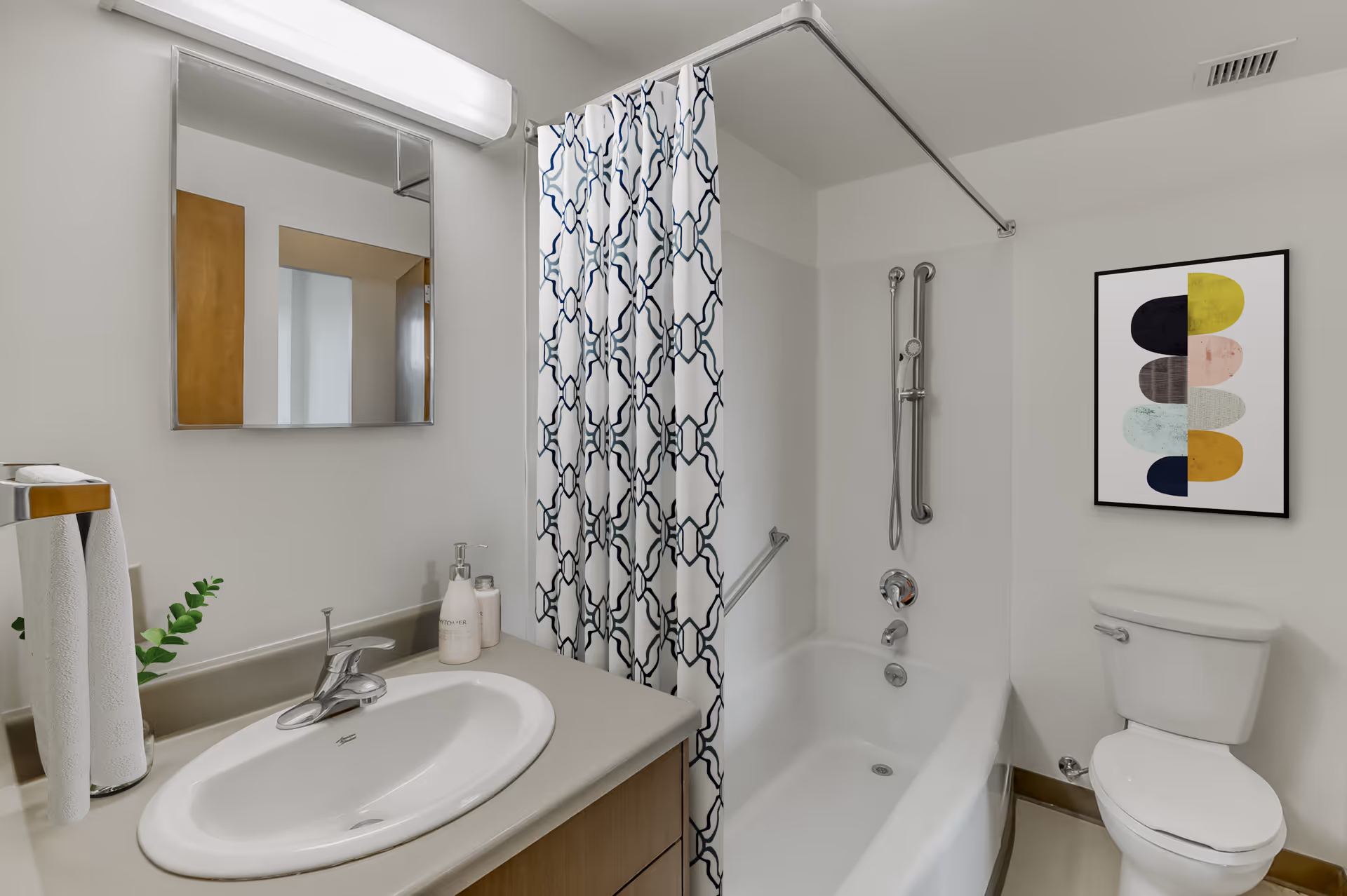 A clean and modern bathroom featuring a white sink with a silver faucet, a mirror with a light above it, a bathtub with a showerhead and a patterned shower curtain, a white toilet, and a colorful abstract framed artwork on the wall.