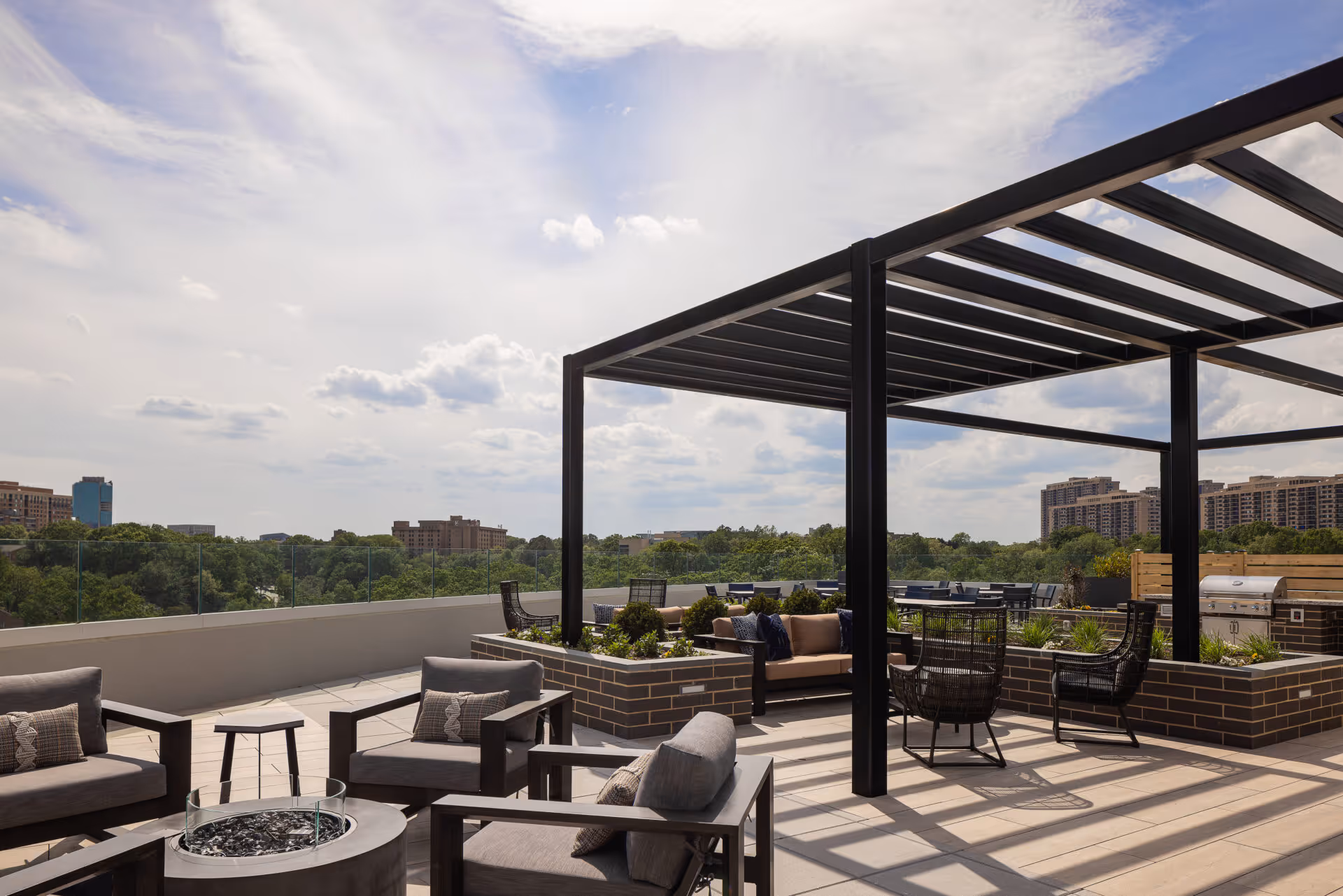 Outdoor rooftop patio area with modern seating including cushioned chairs and sofas, a pergola providing partial shade, planters with greenery, and a grill in the background. The scene overlooks a cityscape with trees and buildings under a partly cloudy sky.