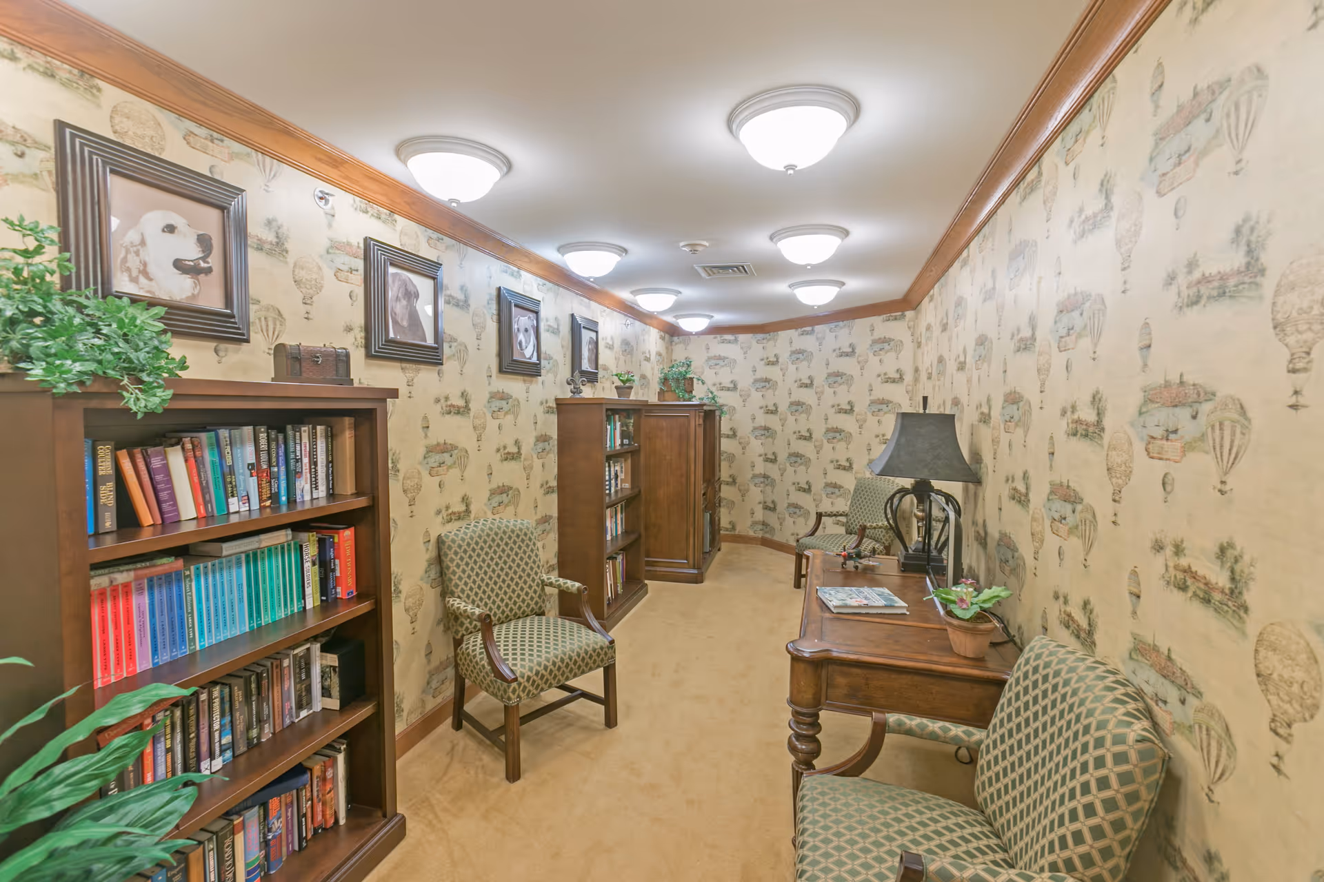 A cozy reading nook in a senior living facility with wallpaper featuring hot air balloons and pastoral scenes. The room has wooden bookshelves filled with books, framed pictures of dogs on the wall, patterned upholstered chairs, a wooden table with a lamp and a small potted plant, and soft overhead lighting.