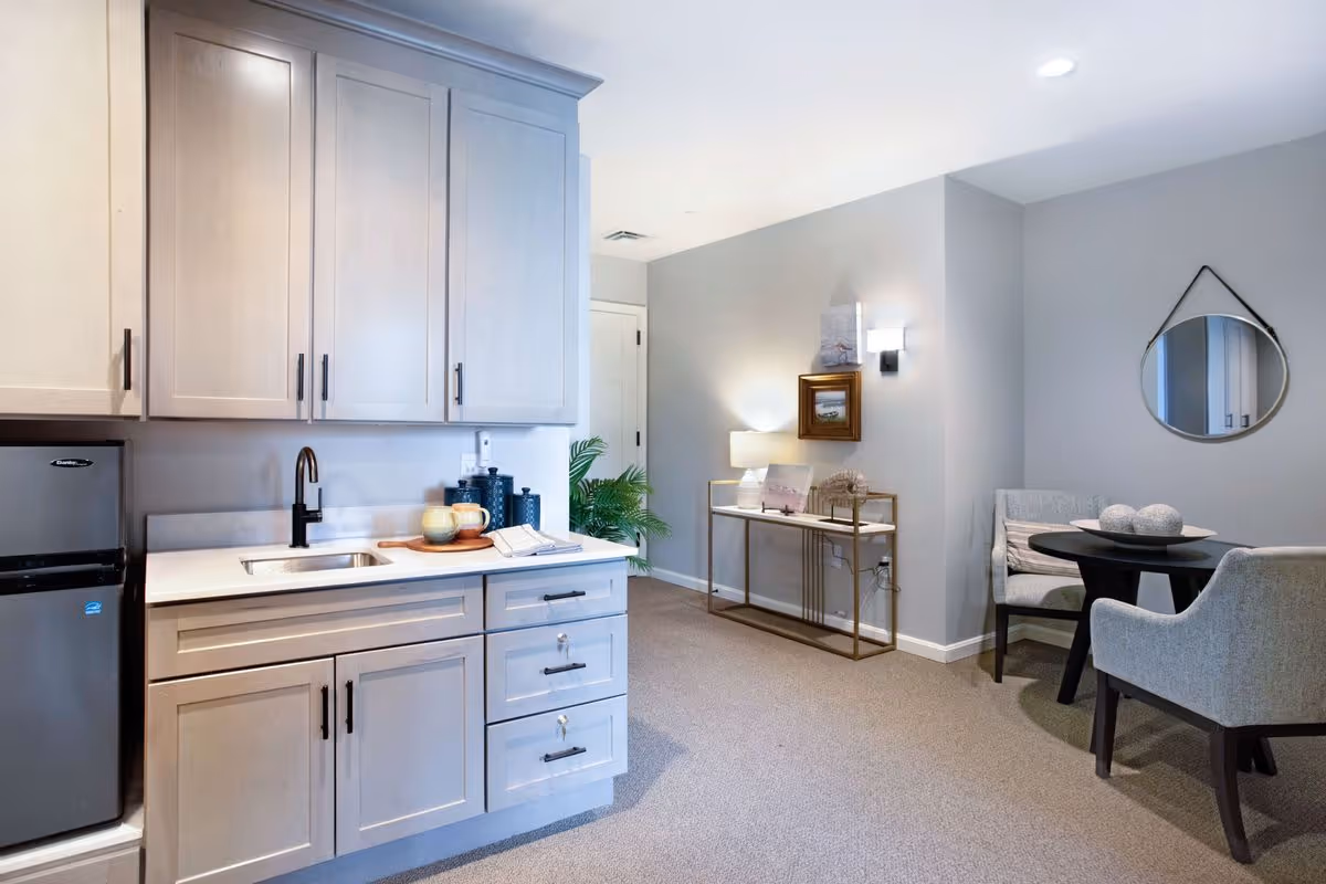 A modern interior space featuring a kitchenette with light gray cabinets, a small stainless steel sink, a black faucet, and a compact refrigerator. To the right, there is a small round black table with two gray upholstered chairs. A console table with a lamp, framed pictures, and decorative items is positioned against the wall, which is painted light gray. A round mirror hangs on the wall above one of the chairs. The room is carpeted and well-lit with ceiling lights and a wall sconce.