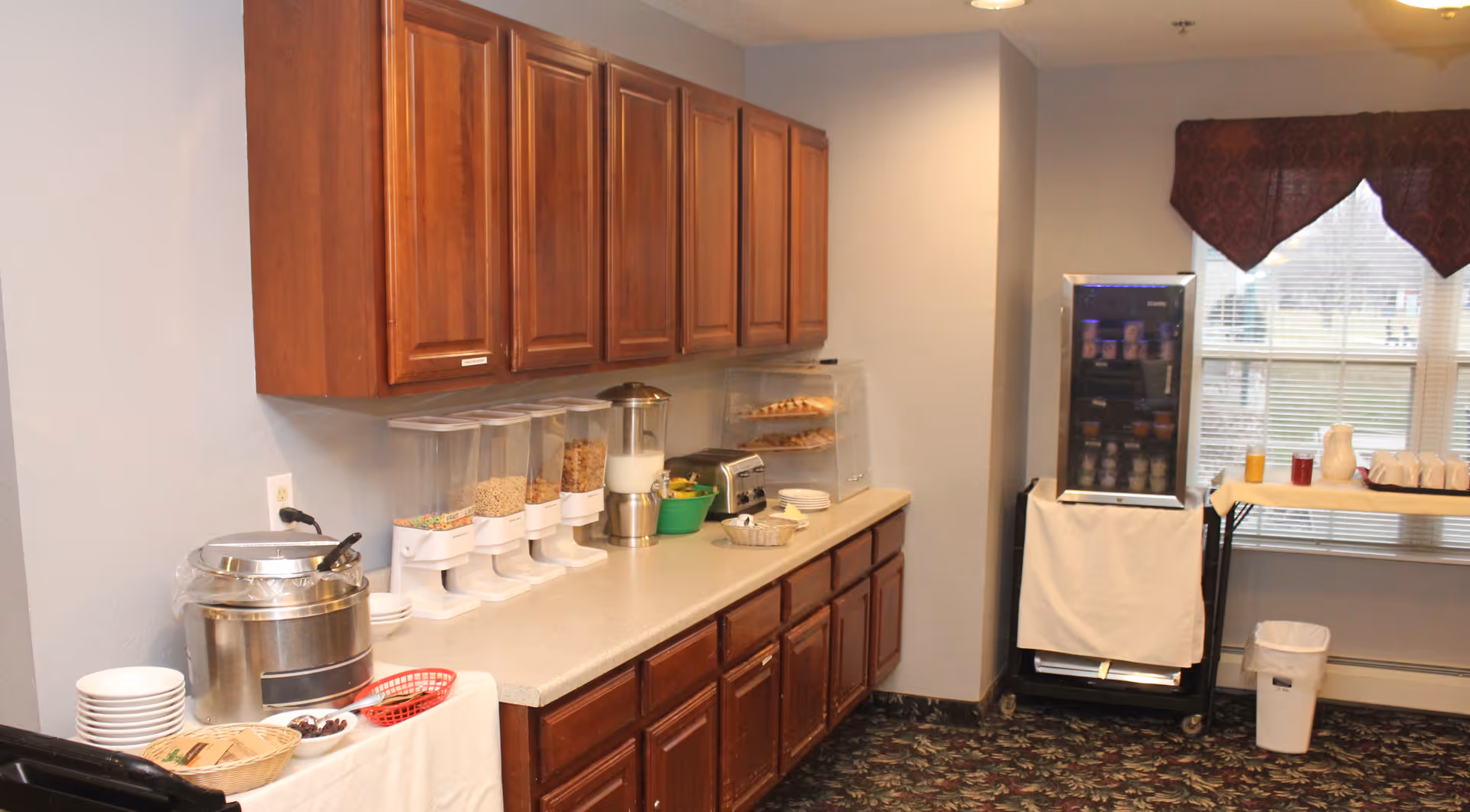 Small buffet/kitchenette area with cereal dispensers, a toaster, coffee urn, cabinets and a refrigerated display by a window.