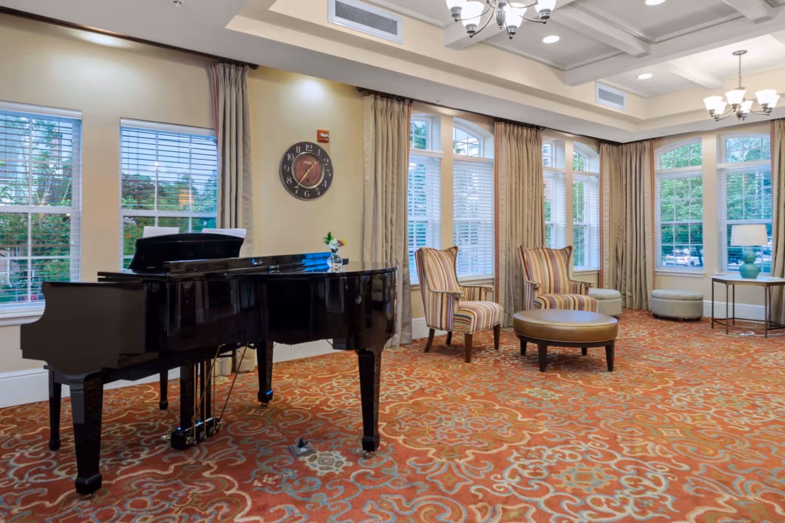 Sunlit communal lounge with a black grand piano, striped armchairs, an ottoman, and large windows.