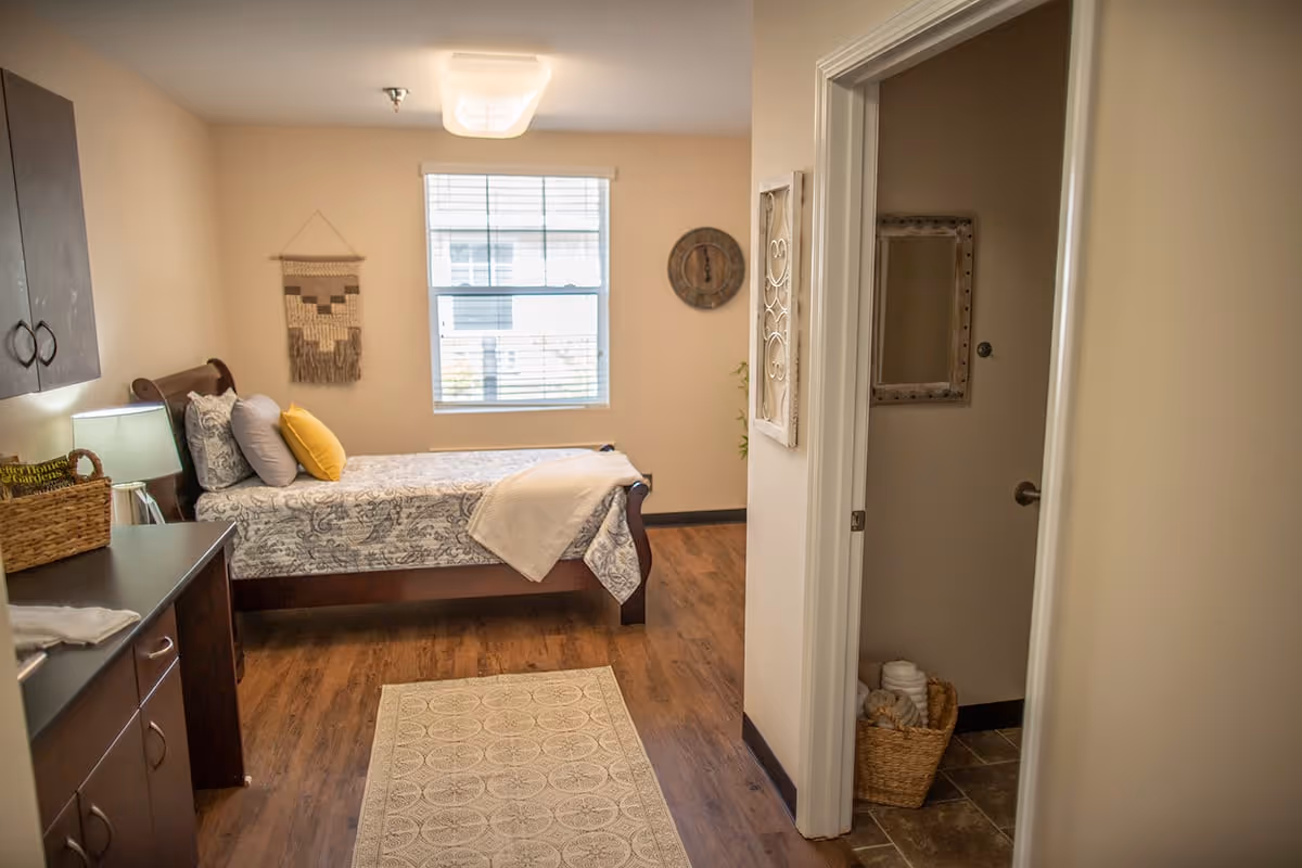 A cozy bedroom with a wooden bed featuring patterned bedding and multiple pillows, including a yellow one. There is a window with blinds letting in natural light, a wall clock, and a decorative wall hanging. To the right, there is an open doorway leading to a bathroom with a basket of towels on the floor. The room has wooden flooring and a small rug in front of the bed.