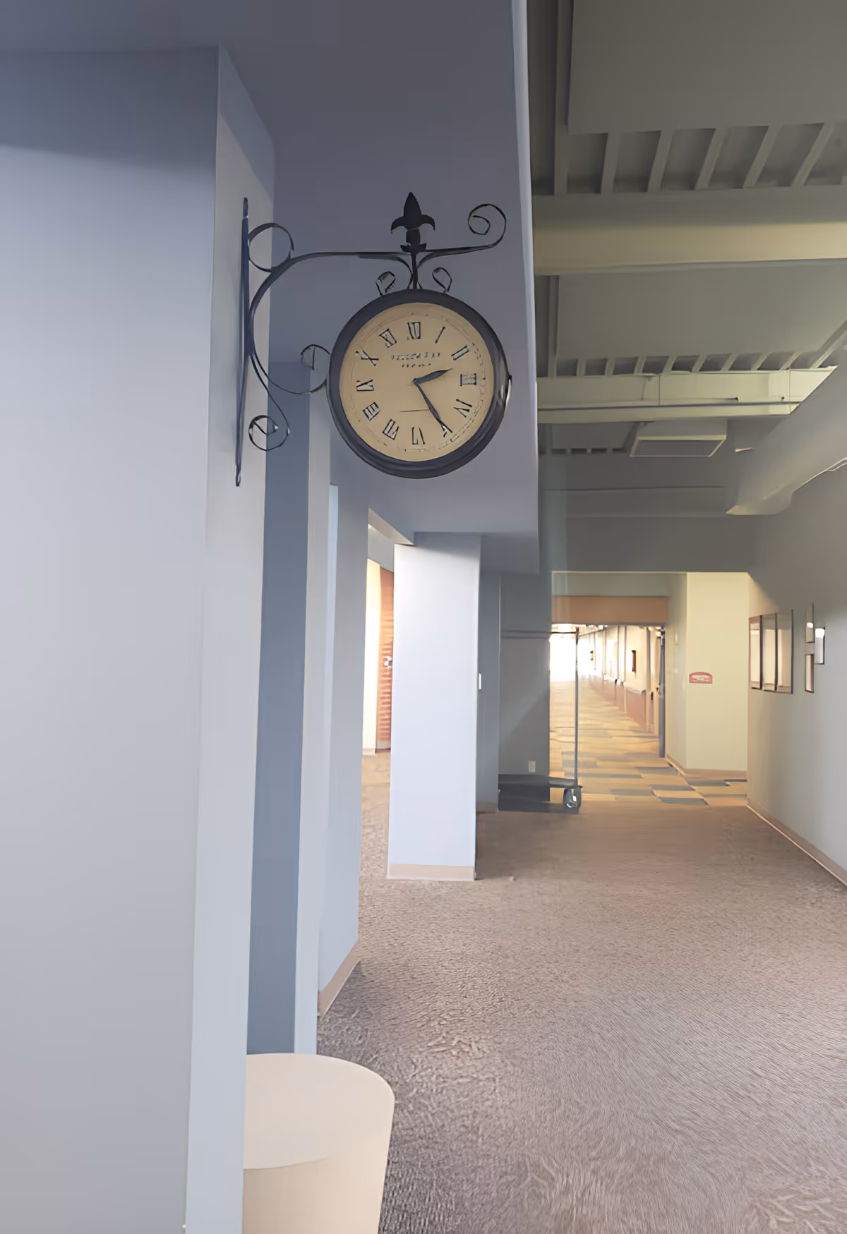 Interior hallway of a facility with a vintage-style wall clock mounted on a decorative metal bracket. The hallway has light blue walls, carpeted floor, and visible ceiling beams. There are framed pictures on the right wall and a glass door at the end of the hallway.