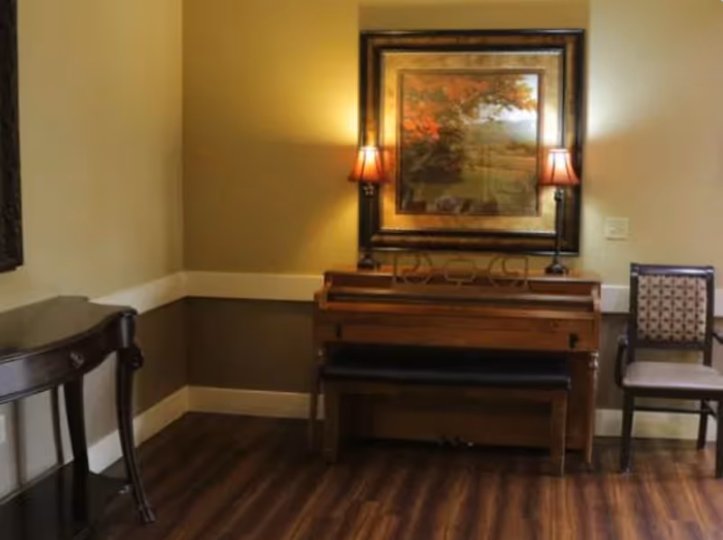 A cozy interior corner featuring a wooden piano with a bench, flanked by two table lamps with warm light. Above the piano hangs a framed painting of a tree with autumn foliage. To the right of the piano is a cushioned chair with a patterned backrest, and to the left is a small dark wooden table. The room has wood flooring and walls painted in two tones with a white chair rail.