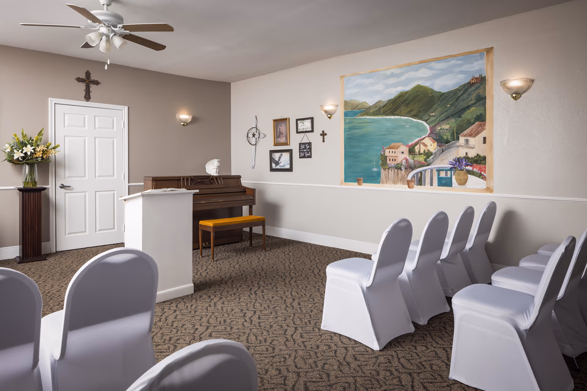 A small chapel or gathering room with rows of white covered chairs facing a wooden piano and a white podium. The walls are decorated with a large mural of a coastal landscape, religious crosses, and framed pictures. A ceiling fan with lights is mounted on the ceiling, and a vase with flowers is placed on a pedestal near a closed white door.