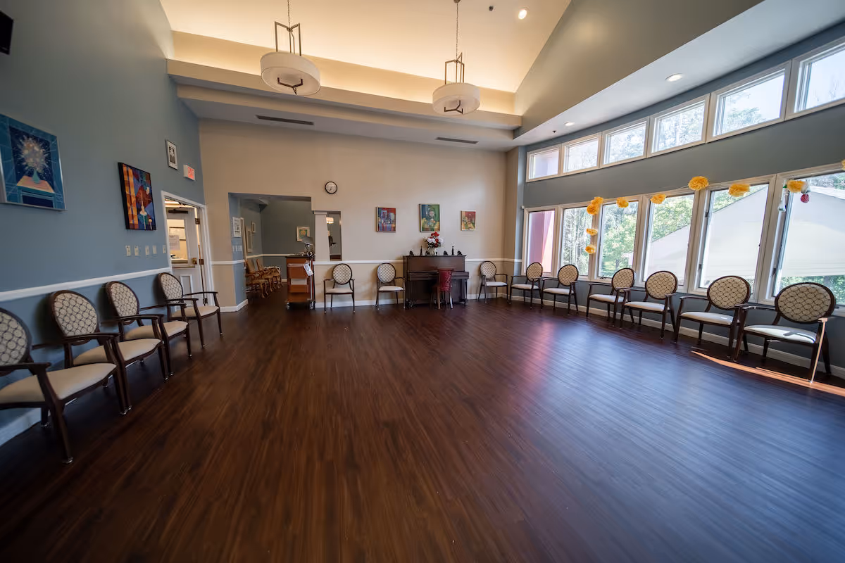 A spacious room with wooden flooring and light blue walls, featuring a row of cushioned chairs with patterned upholstery arranged along two walls. Large windows on one side let in natural light, and there are two modern ceiling light fixtures. The room also contains a piano with a flower arrangement on top and several framed artworks on the walls.
