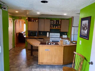 Interior view of a kitchen with green walls, wooden cabinets, a stove, microwave, and a kitchen island with a sink. There is a framed picture on the right wall and a wooden chair partially visible in the foreground. A hallway leads to another room in the background.