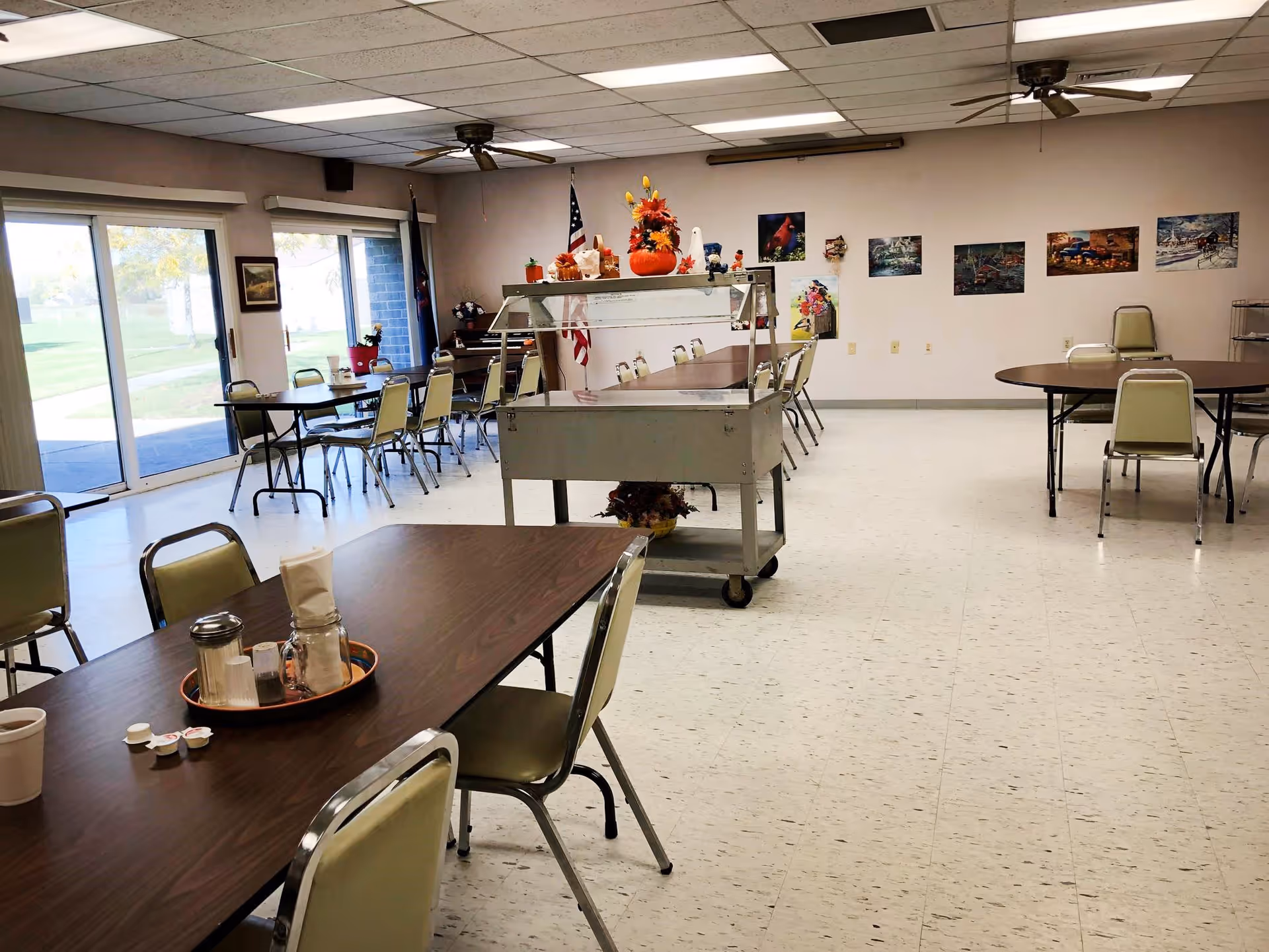 Spacious community dining room with multiple tables and chairs, a serving cart in the center, and sliding glass doors to the outside.