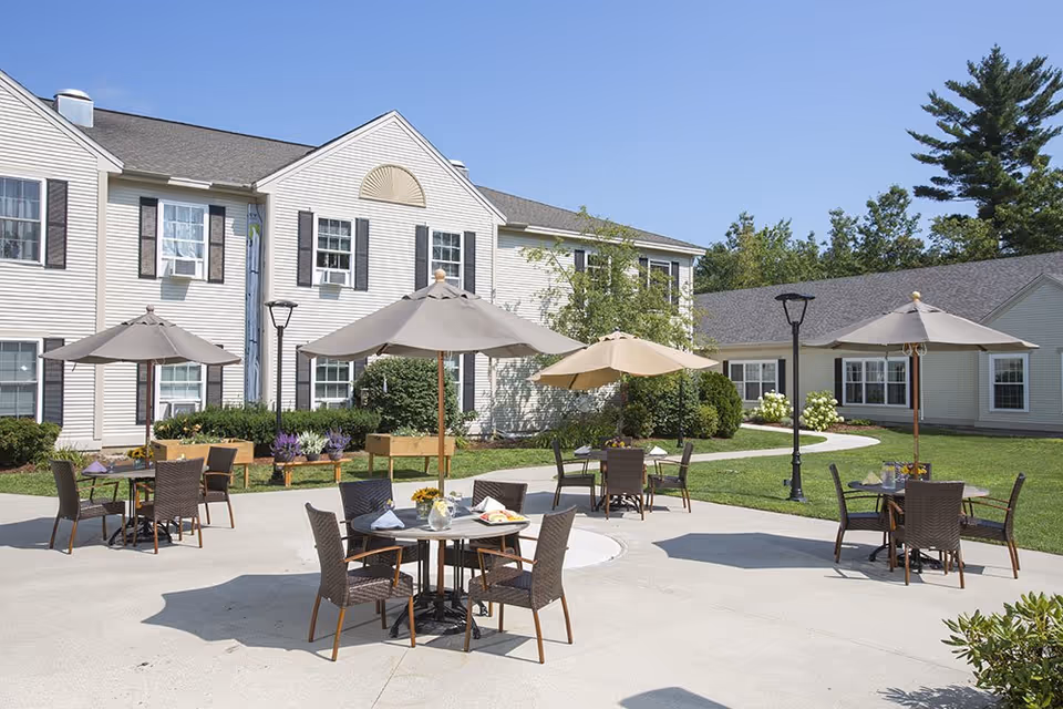 Outdoor patio area at Benchmark Senior Living at Nashua Crossings with several round tables and chairs under large umbrellas on a concrete surface, surrounded by green grass and shrubs, with a two-story beige building in the background under a clear blue sky.