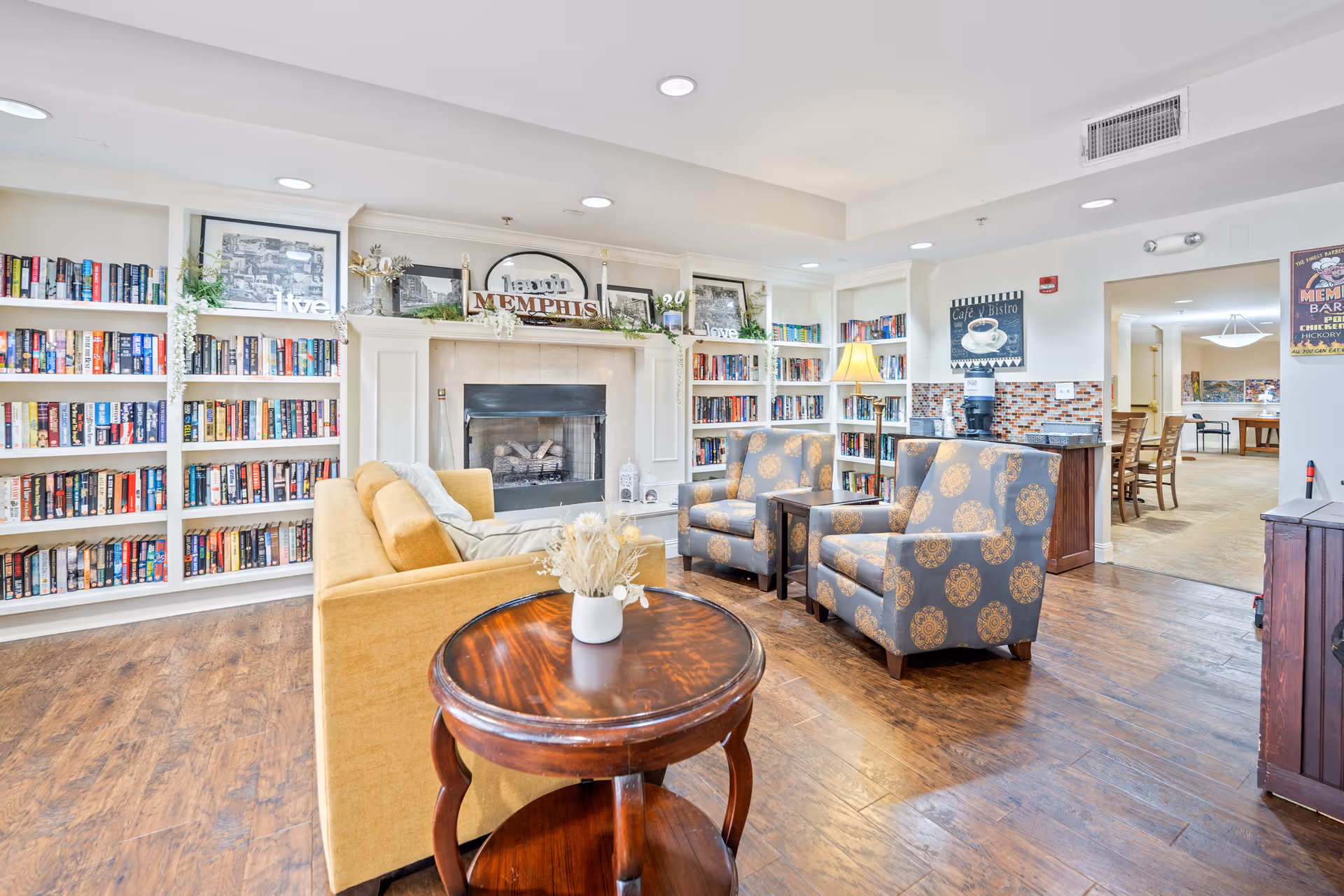 A cozy living room area in a senior living facility with a yellow sofa, two patterned armchairs, a round wooden coffee table with a vase of dried flowers, and built-in bookshelves filled with books surrounding a fireplace. The room has wooden flooring and recessed ceiling lights, with a view into an adjacent dining area.