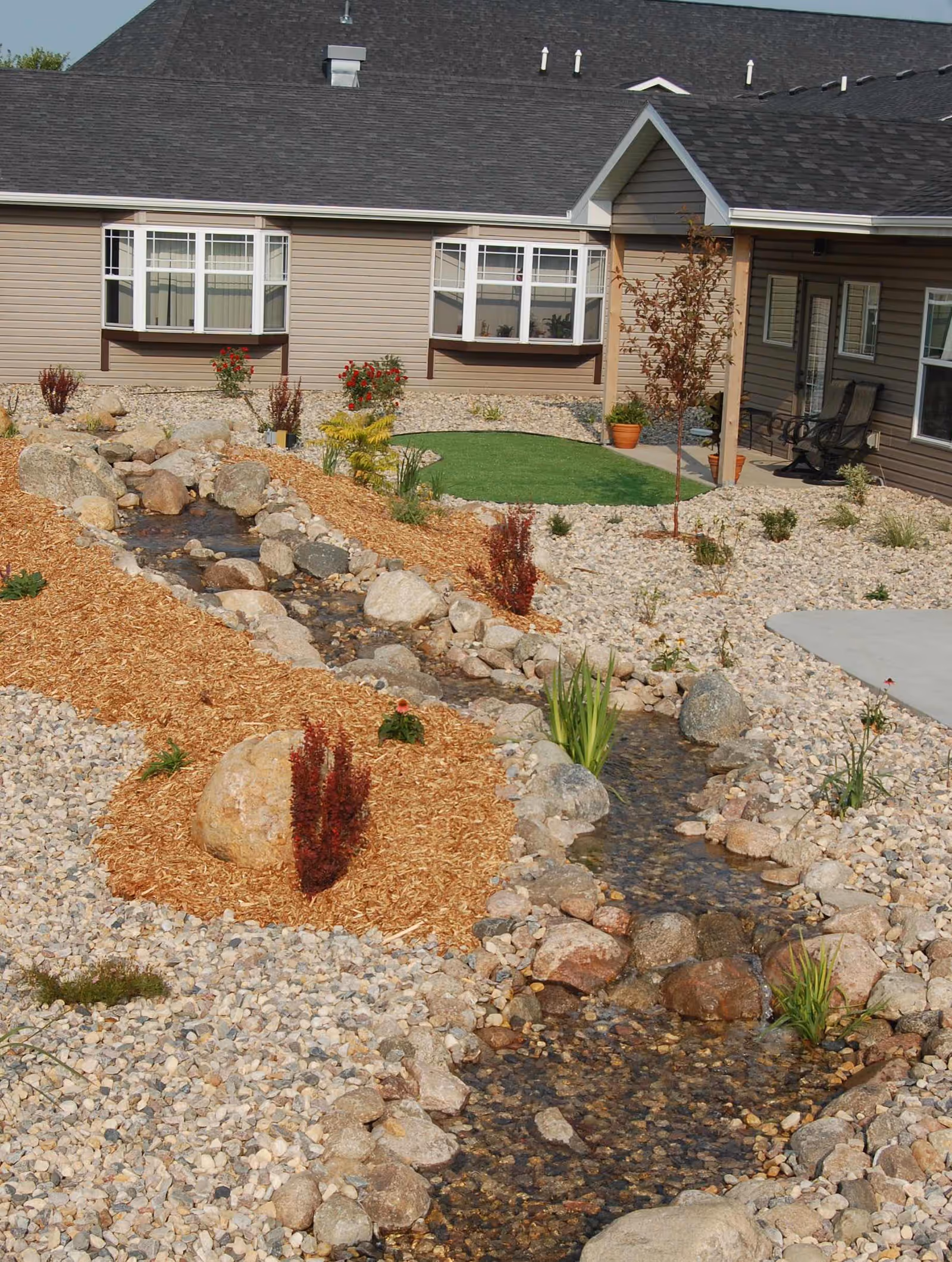A landscaped outdoor area at StoneyBrook Suites Assisted Living featuring a small artificial stream bordered by rocks and mulch, various plants and shrubs, a patch of artificial grass, and a building with beige siding and multiple windows in the background.
