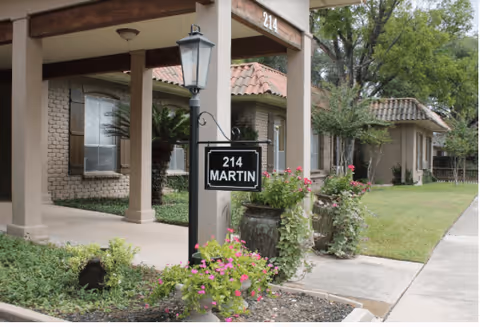 Exterior view of a retirement residence building with beige brick walls and a tiled roof. There is a covered walkway supported by columns, a black lamp post with a sign reading '214 MARTIN', and landscaped greenery including flowering plants and shrubs.