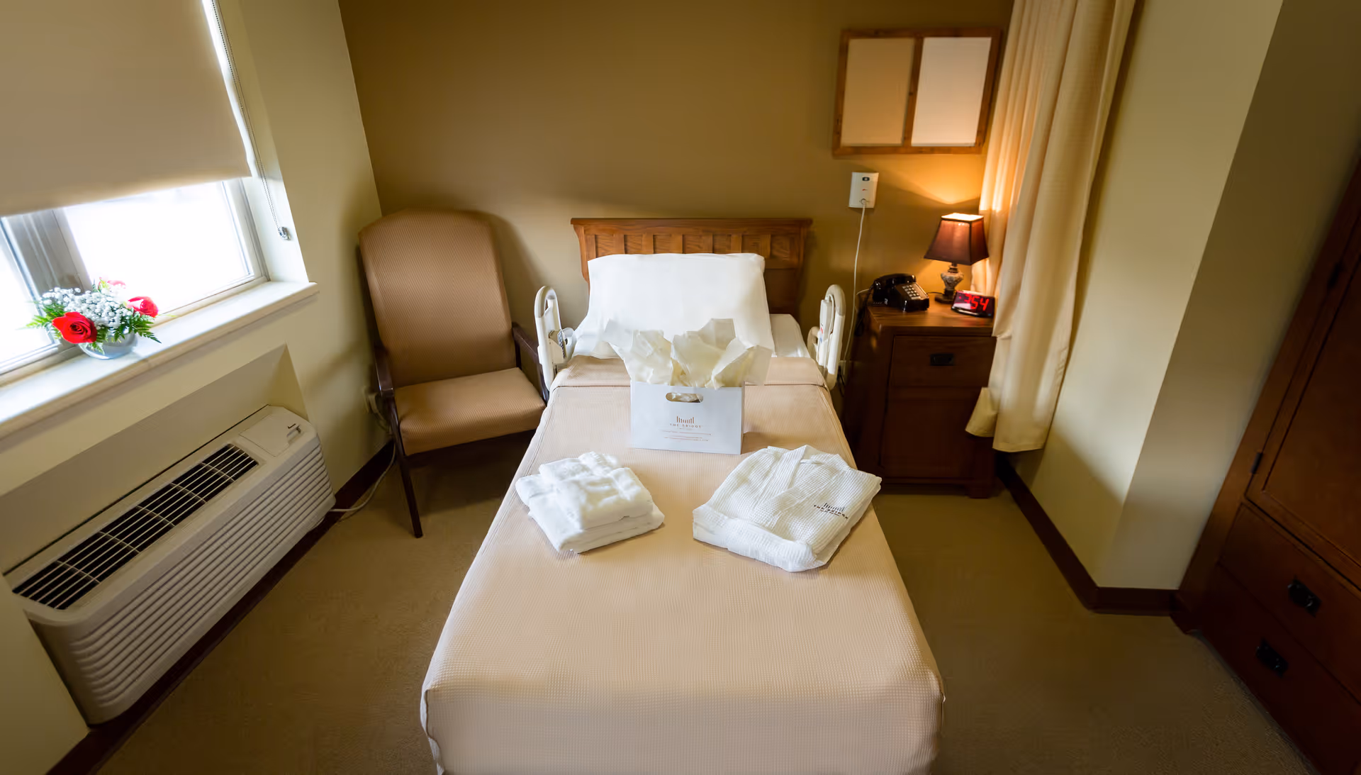 A small, neatly arranged bedroom in a senior living facility with a single bed covered in beige bedding. On the bed are folded white towels and a gift bag. To the left of the bed is a beige armchair next to a window with a flower arrangement on the windowsill. Below the window is an air conditioning unit. To the right of the bed is a wooden nightstand with a lamp, a telephone, and a digital clock showing 3:41. Beige curtains hang beside the nightstand, and a wooden wardrobe is visible on the far right.