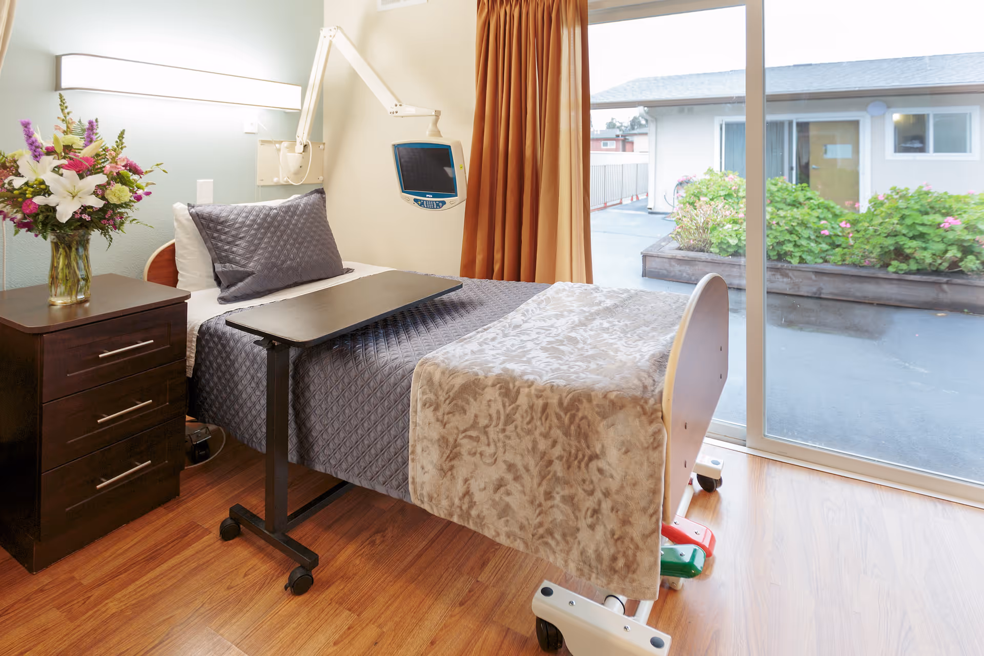 A tidy senior bedroom featuring an adjustable hospital-style bed with an overbed table, a nightstand with flowers, and sliding glass doors overlooking a courtyard.