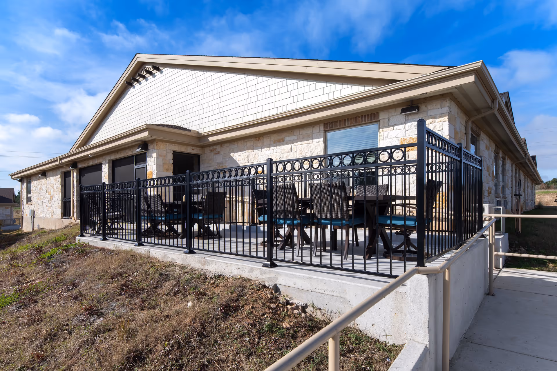 Outdoor patio area of a stone building with a black metal fence surrounding a seating area with tables and chairs. The sky is blue with some clouds.