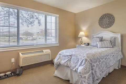 A cozy bedroom with a single bed covered in a patterned white and gray bedspread, a white headboard, and several pillows. Next to the bed is a white nightstand with a lamp and a small clock. A large window with white blinds lets in natural light, and below the window is an air conditioning unit. The walls are painted beige, and a decorative round wall piece hangs above the bed.