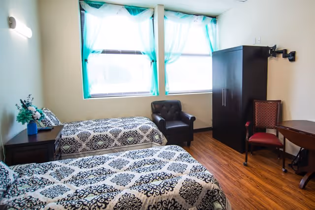 A bright bedroom with two twin beds covered in black and white patterned bedspreads. There is a small dark wooden nightstand with a vase of flowers between the beds. A black armchair is placed near two large windows with sheer turquoise curtains. The room also contains a dark wardrobe, a red upholstered chair, and a wooden table on a wood floor.
