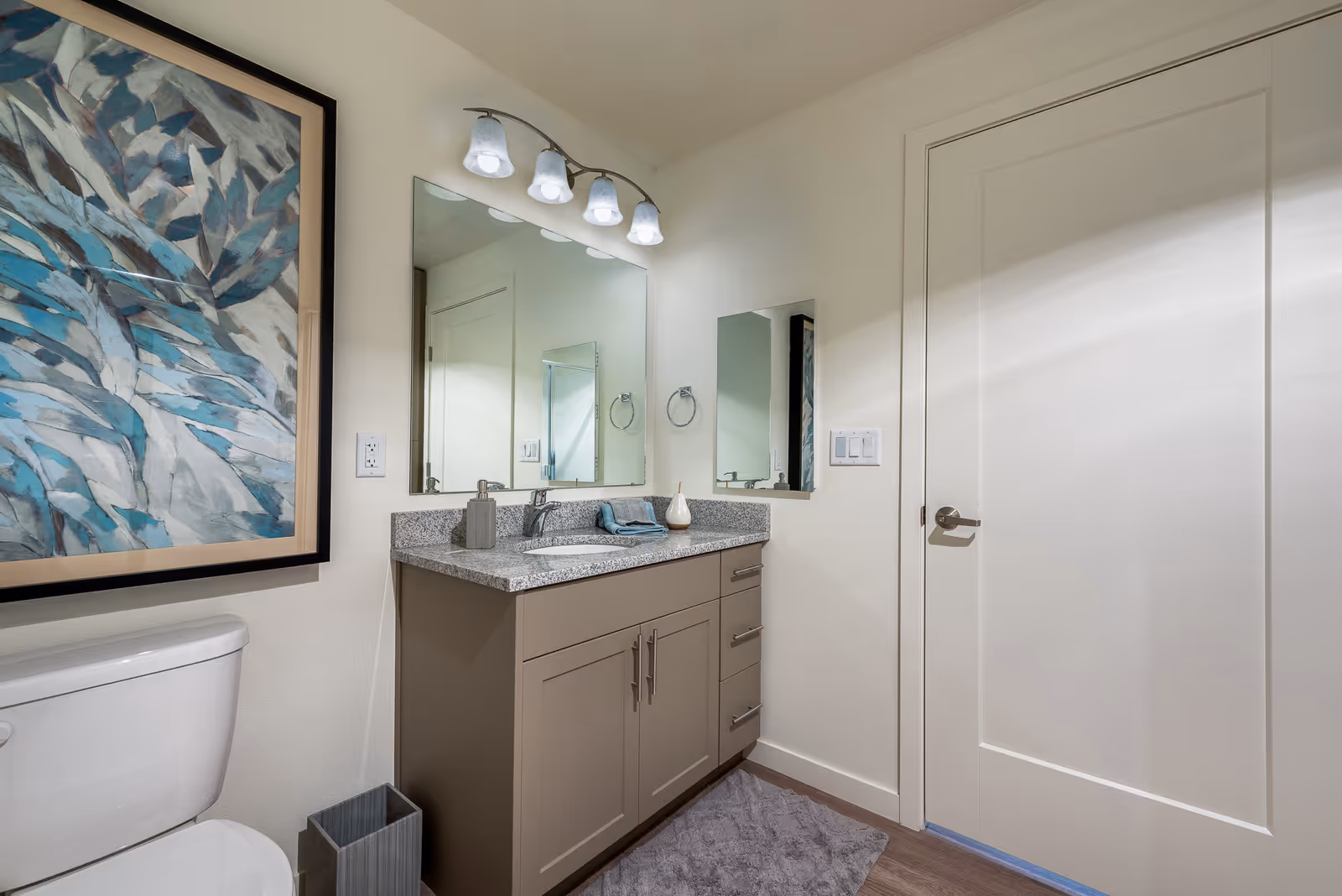 A modern bathroom featuring a white toilet, a gray vanity with a granite countertop, a large mirror with four light fixtures above it, a small rectangular mirror on the adjacent wall, a gray hand towel, a soap dispenser, a small decorative item on the countertop, a gray waste bin, a textured gray bath mat on the wooden floor, and a large framed abstract artwork with blue and gray tones on the wall.