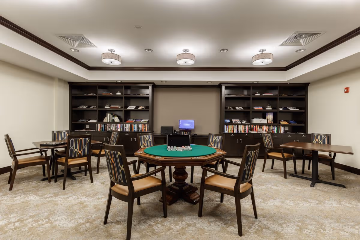 A well-lit room with a central round table covered with a green felt surface and surrounded by six chairs with patterned backs and cushioned seats. There are two rectangular tables with similar chairs on either side of the room. The back wall features two large dark wood bookshelves filled with books and decorative items, with a small computer monitor placed in the center between the shelves. The ceiling has recessed lighting and three modern circular light fixtures.