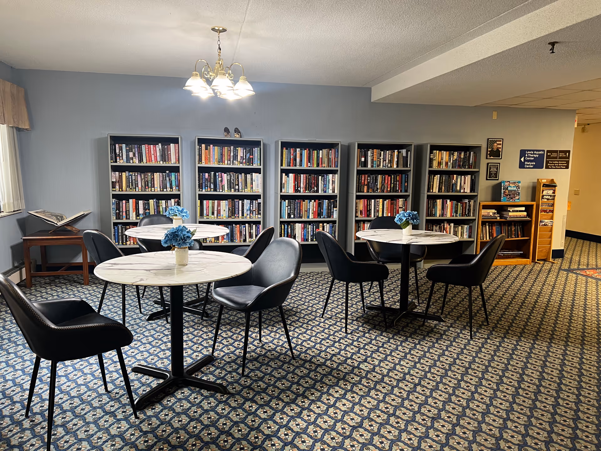 A cozy reading area in Myers Apartments Independent Living featuring several round tables with black chairs around them. Each table has a small vase with blue flowers. Behind the tables are multiple bookshelves filled with books against a light blue wall. The floor is covered with a patterned carpet, and a chandelier with multiple lights hangs from the ceiling.