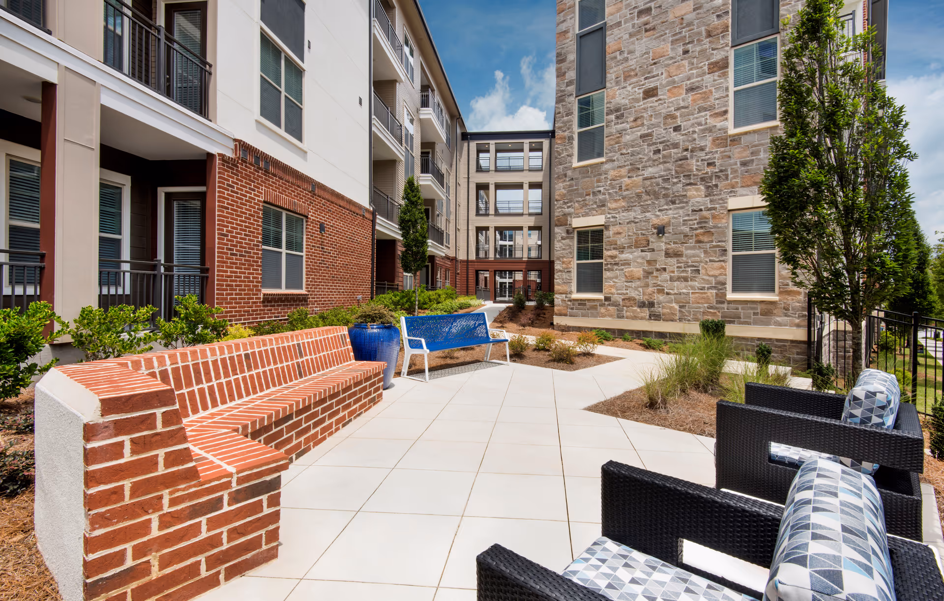 Outdoor courtyard area of a senior living facility with brick and stone buildings surrounding a tiled patio. The patio features a built-in brick bench, a blue metal bench, and black wicker chairs with patterned cushions. There are small landscaped areas with shrubs and trees, and a blue sky with some clouds overhead.