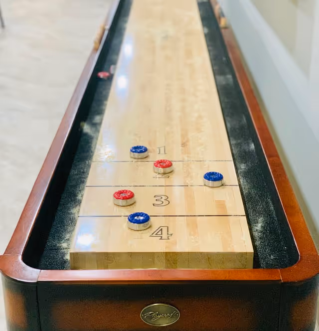 A shuffleboard table with red and blue pucks positioned on the numbered scoring zones, set in an indoor space with a wooden frame and a light-colored playing surface.