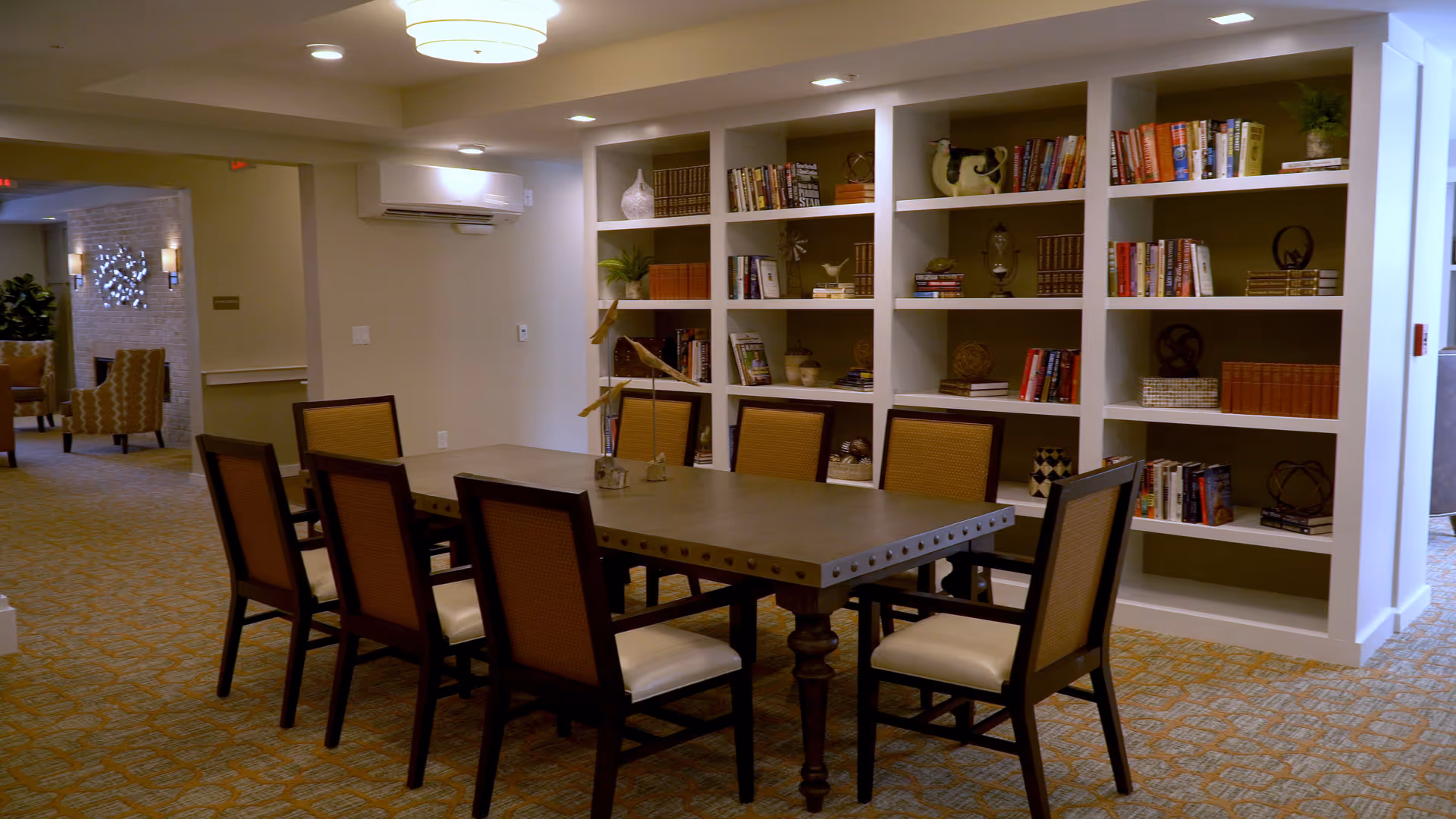 A well-lit interior room with a large rectangular table surrounded by eight chairs. Behind the table is a built-in bookshelf filled with books and decorative items. The room has carpeted flooring and neutral-colored walls, with additional seating visible in the background hallway.