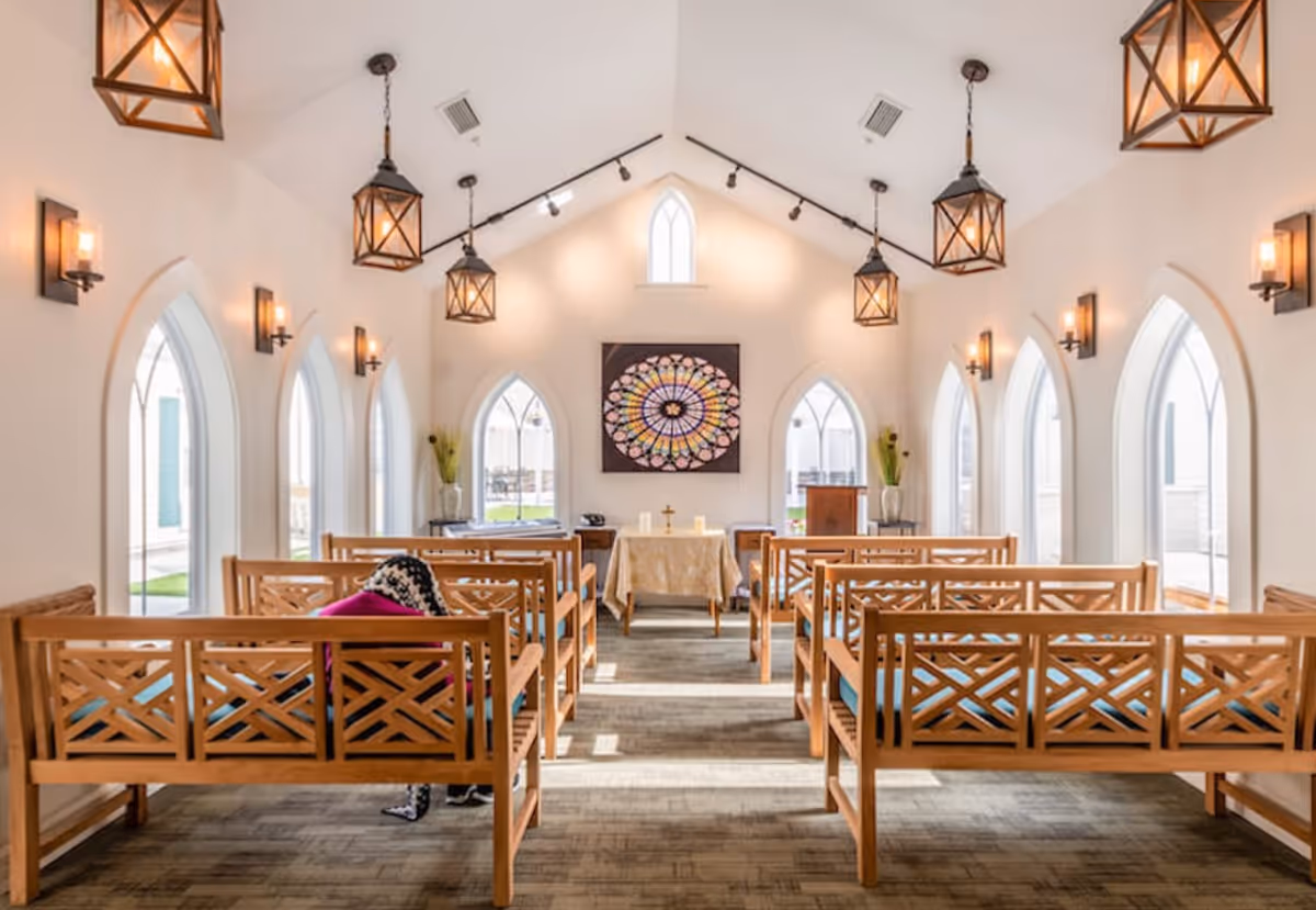 Interior of a small chapel or meditation room with wooden benches arranged in rows facing an altar. The room has high vaulted ceilings with hanging lantern-style lights and arched windows letting in natural light. A colorful circular stained glass artwork is mounted on the wall behind the altar.