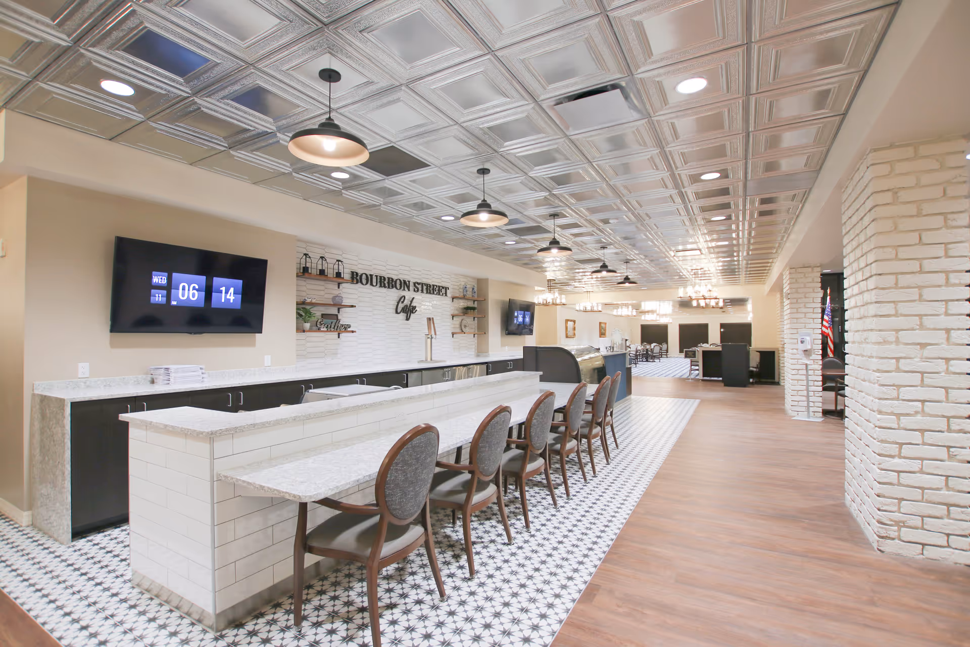 Long café-style dining area with a tiled service counter, a row of chairs, patterned floor tiles, and a decorative tin ceiling under pendant lights.
