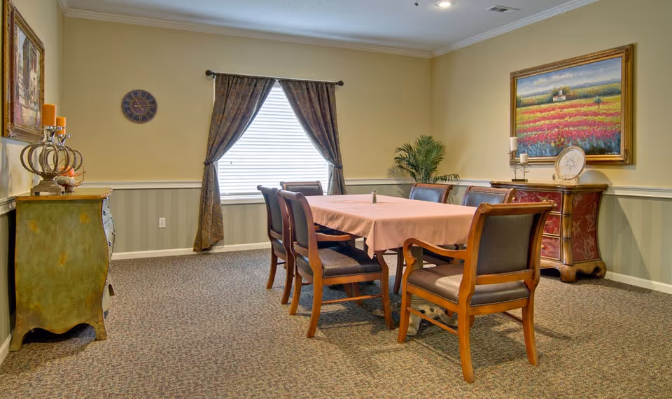 A dining room with a rectangular table covered with a pink tablecloth surrounded by six wooden chairs with cushioned seats. The room has beige walls with a chair rail and patterned carpet. There are two decorative dressers, one on each side of the room, with candles and artwork above them. A window with dark curtains is centered on the far wall, and a potted plant is placed near the right dresser.