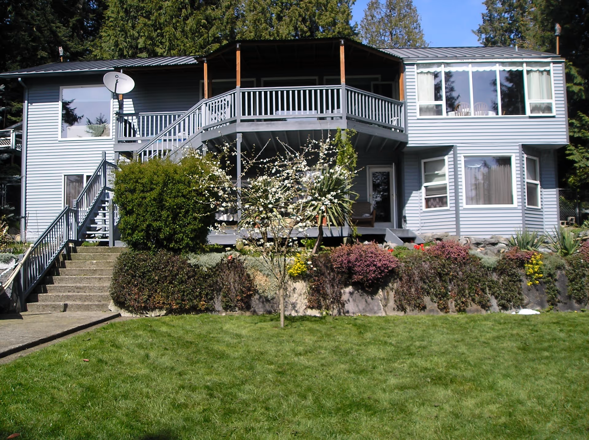 Exterior view of a two-story gray house with a balcony, stairs leading up to the entrance, and a well-maintained garden with bushes, flowers, and a small tree in front. The house is surrounded by tall trees and has large windows on both floors.
