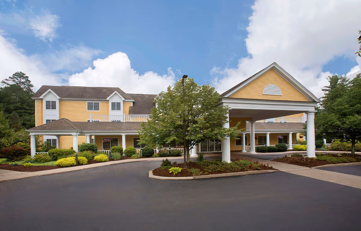 Exterior view of a yellow two-story senior living facility building with white columns and a covered entrance driveway. The building is surrounded by landscaped greenery and trees under a partly cloudy sky.