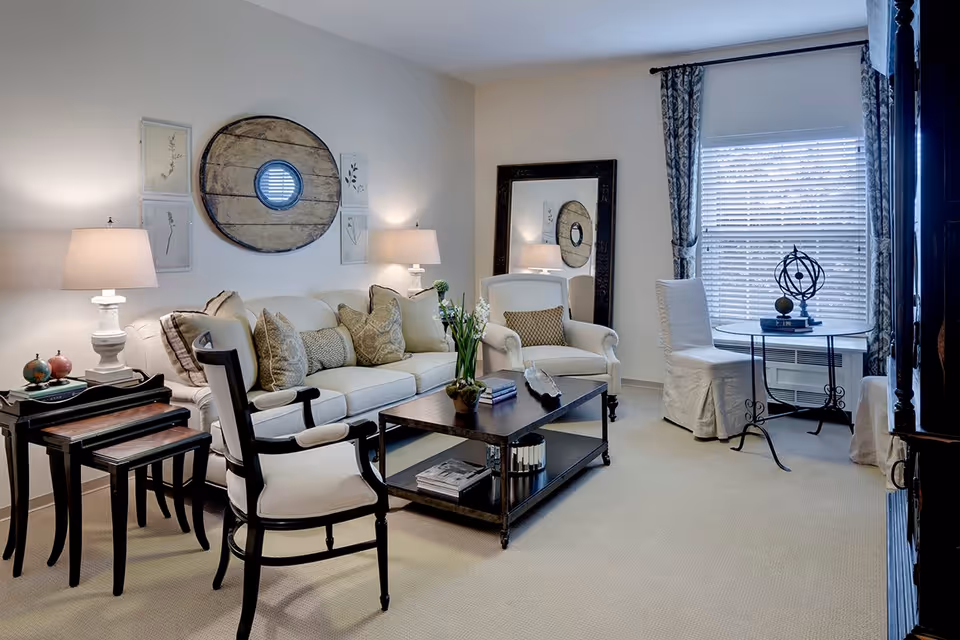 A cozy living room with a white sofa adorned with multiple decorative pillows, two white armchairs, a dark wooden coffee table with books and a plant, and a set of nested tables with two lamps. The walls are decorated with framed artwork and a large round wooden piece. A large mirror leans against the wall near a window with white blinds and patterned curtains. A small round table with two white slipcovered chairs is positioned by the window, featuring a decorative globe and books.