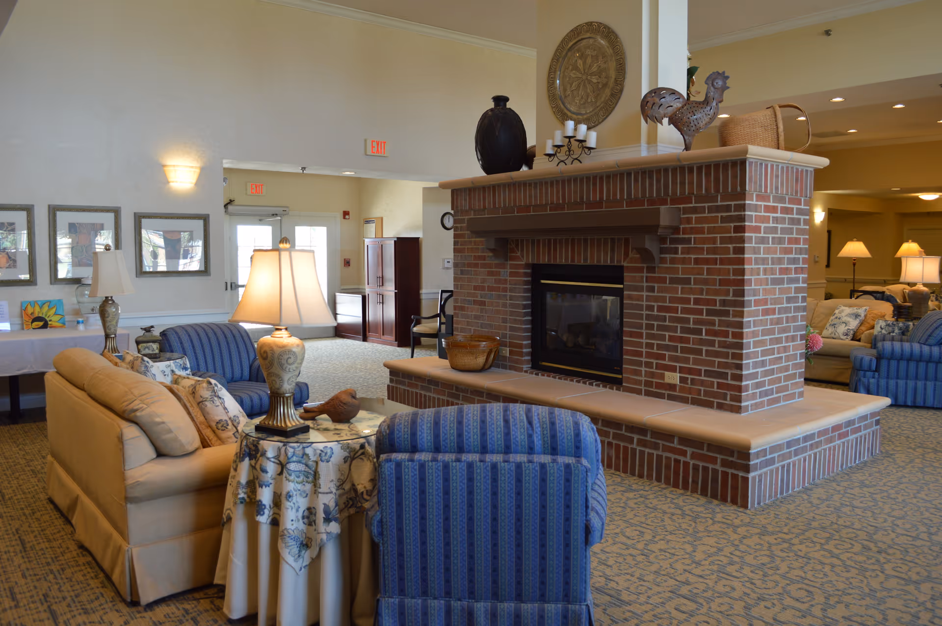 A cozy senior living facility common area with a large brick fireplace in the center. Surrounding the fireplace are comfortable upholstered chairs and sofas in beige and blue tones. A small round table with a floral tablecloth and a decorative lamp sits between the seating. The room is warmly lit with wall sconces and table lamps, and there are framed pictures on the wall. The space has a welcoming and homey atmosphere.