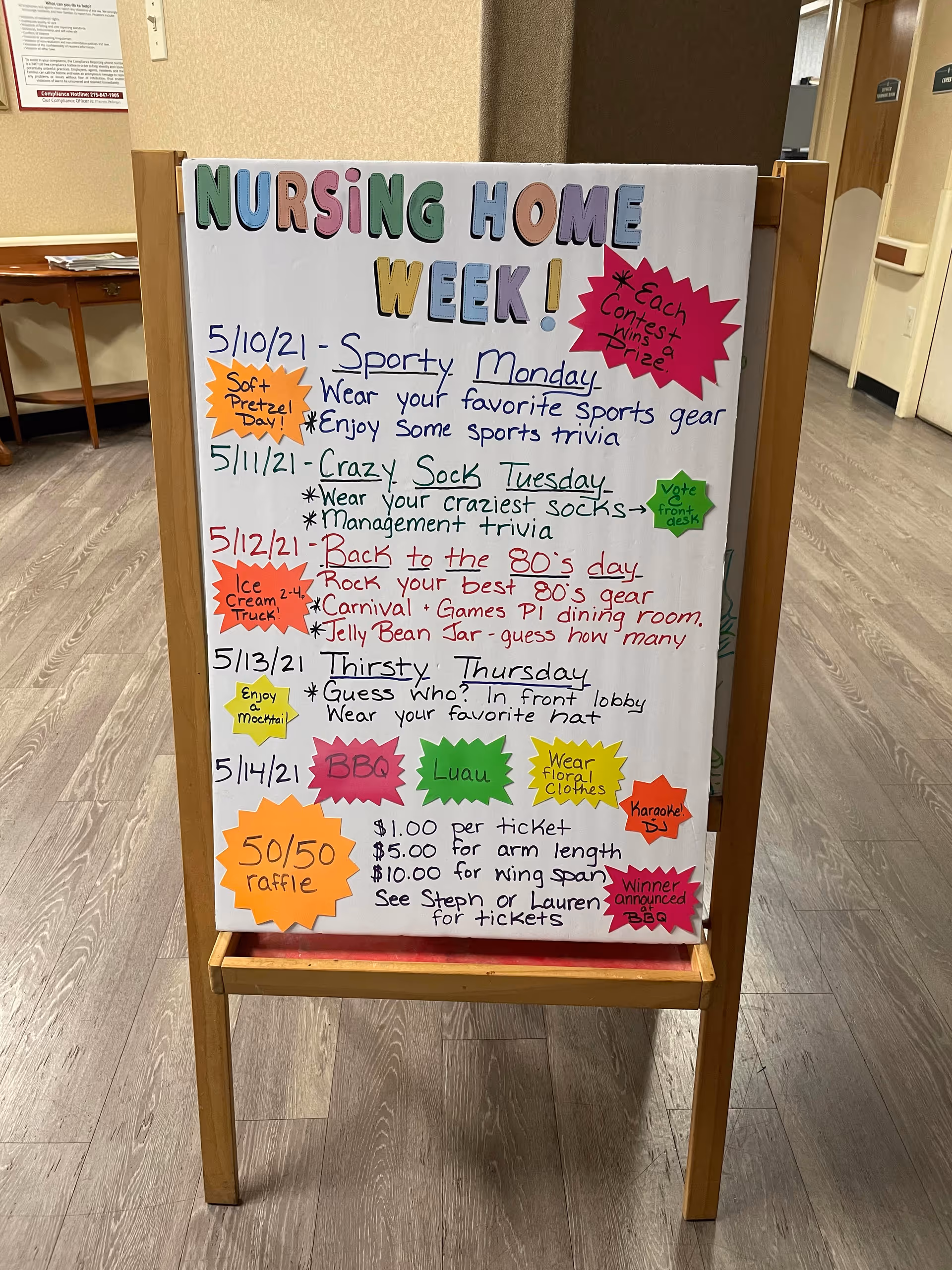 A colorful whiteboard sign on a wooden stand in a hallway, detailing activities for Nursing Home Week with dates and event descriptions including Sporty Monday, Crazy Sock Tuesday, Back to the 80's day, Thirsty Thursday, and a BBQ Luau with a 50/50 raffle. The sign is decorated with various colorful paper cutouts highlighting different events and notes.